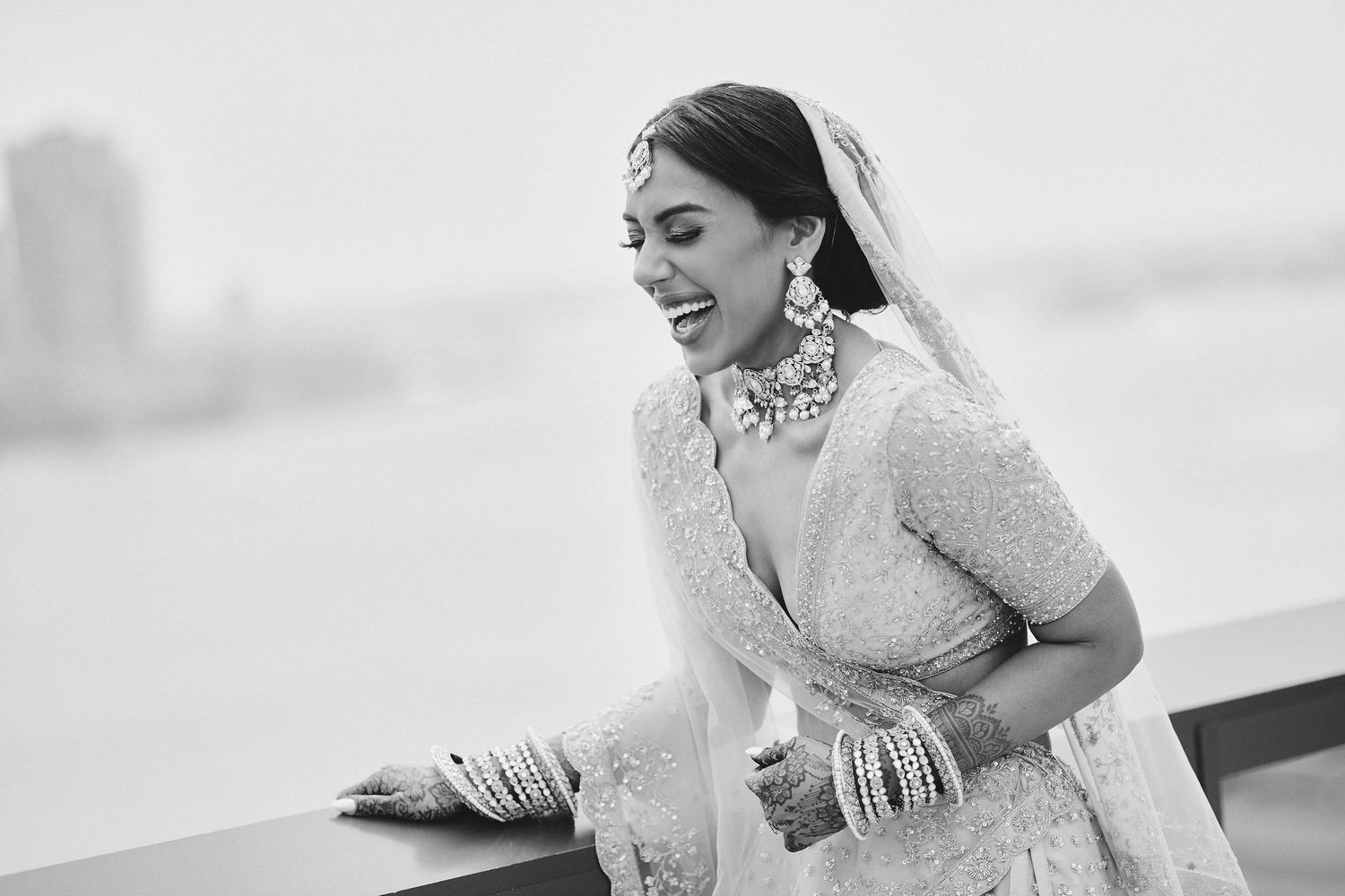 A joyful bride in traditional Indian wedding attire laughs while leaning against a railing.