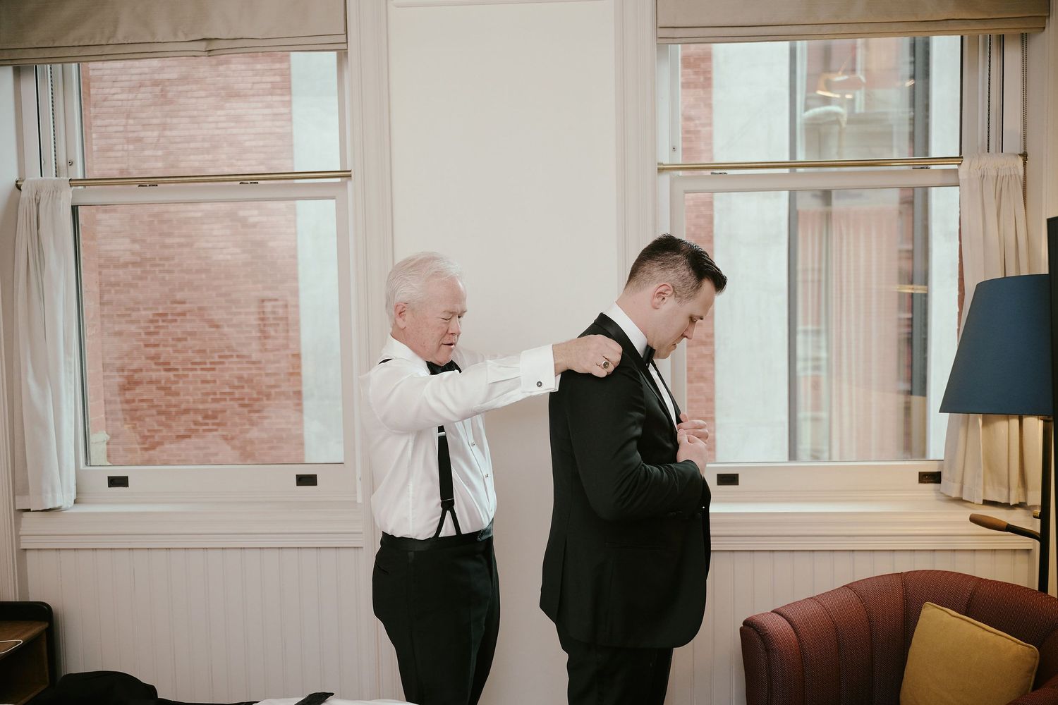 Two people in formal attire help each other get ready in a bright room with white walls and windows.
