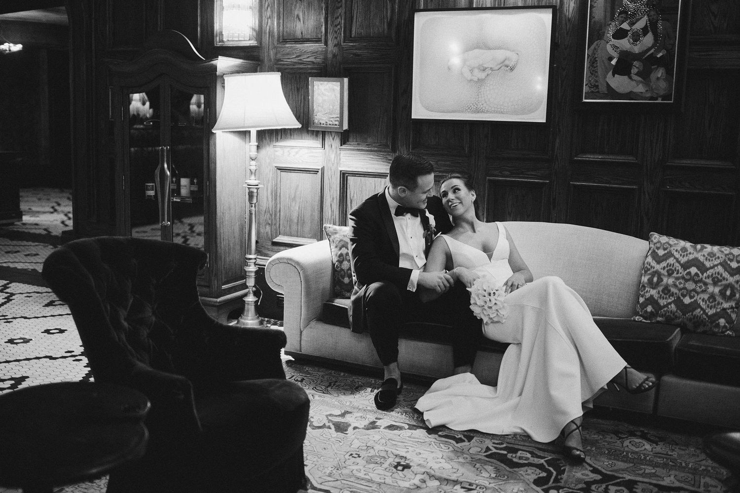 A romantic black and white scene of a couple in formal attire relaxing together on a vintage couch in a dimly lit room.