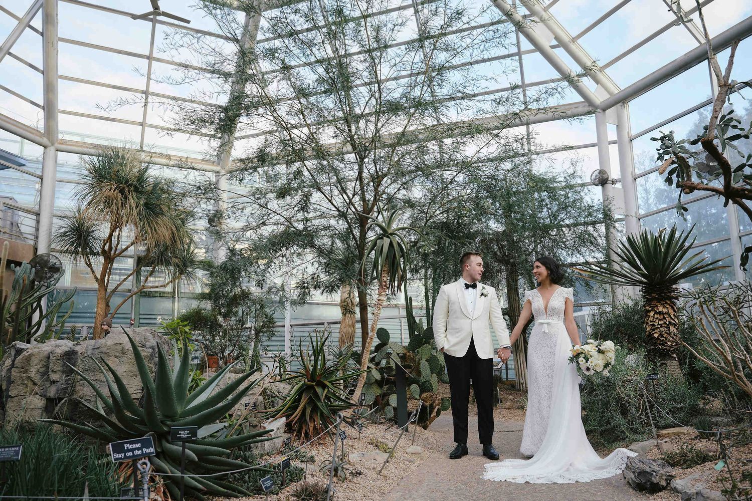 A couple stands in a greenhouse wedding venue surrounded by desert plants and tropical foliage under a glass ceiling.