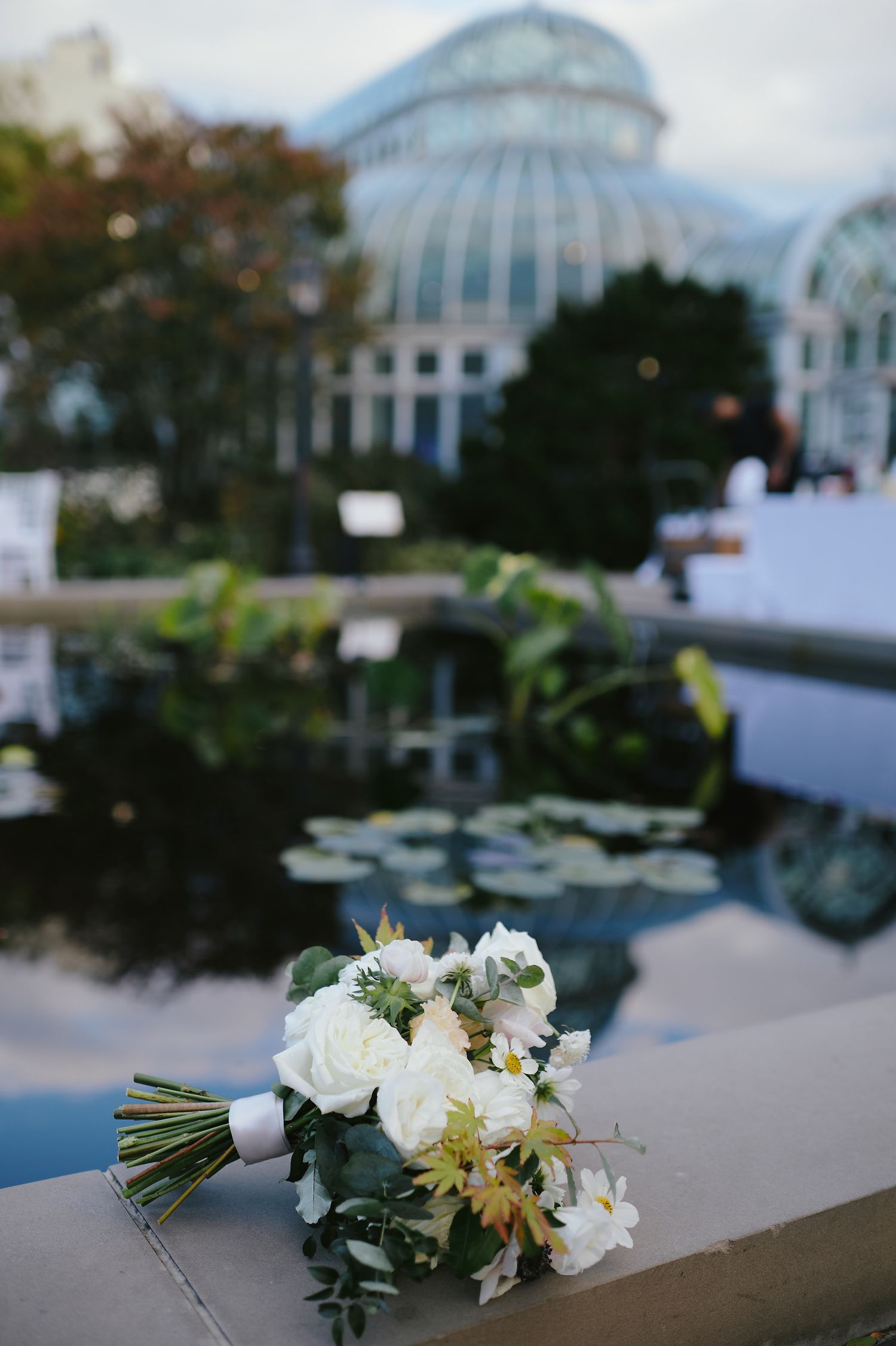 A white wedding bouquet rests on a ledge with a glass conservatory greenhouse and lily pond in the background.
