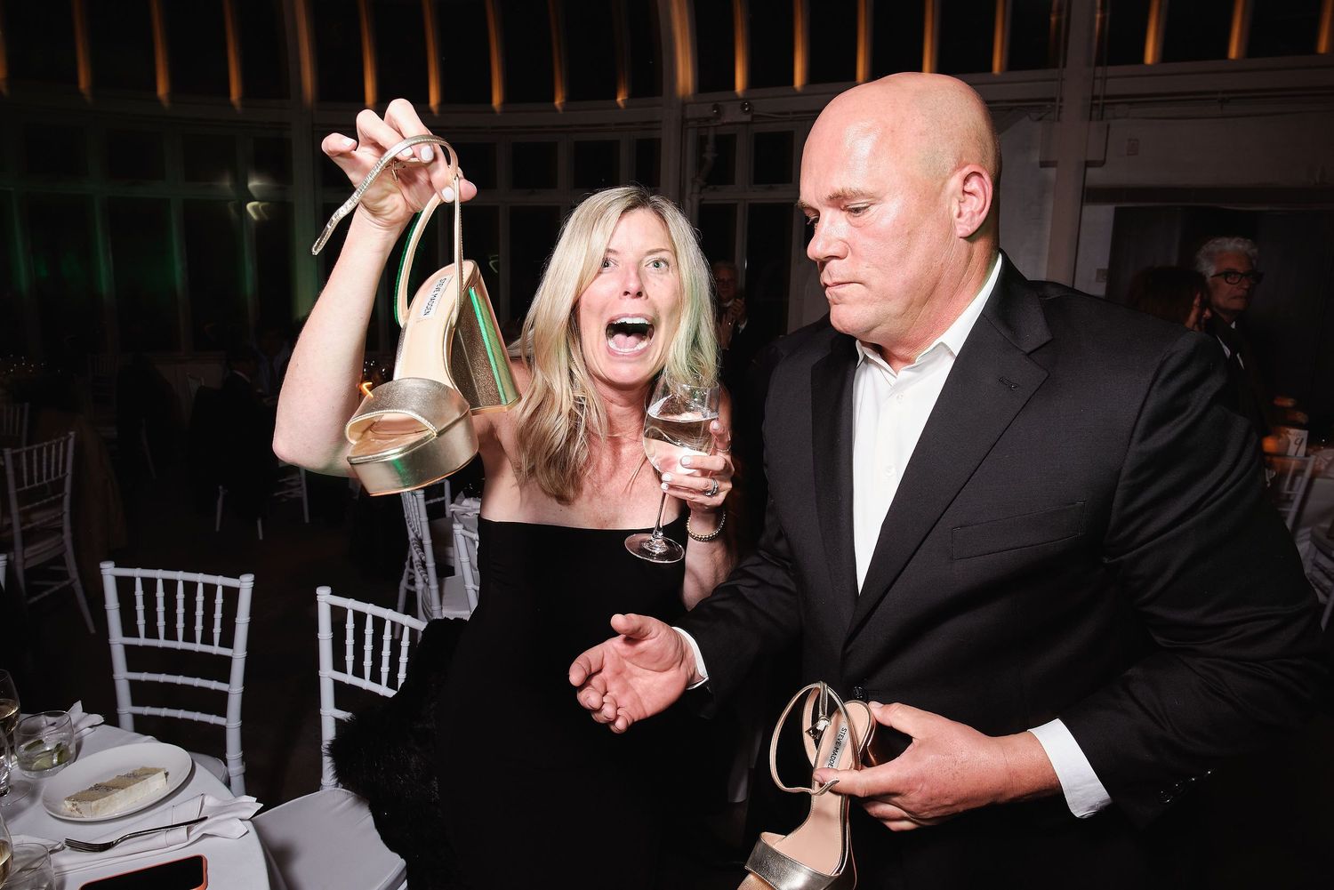 Two people in formal evening attire appear animated and lively while holding drinks at an elegant dining event.