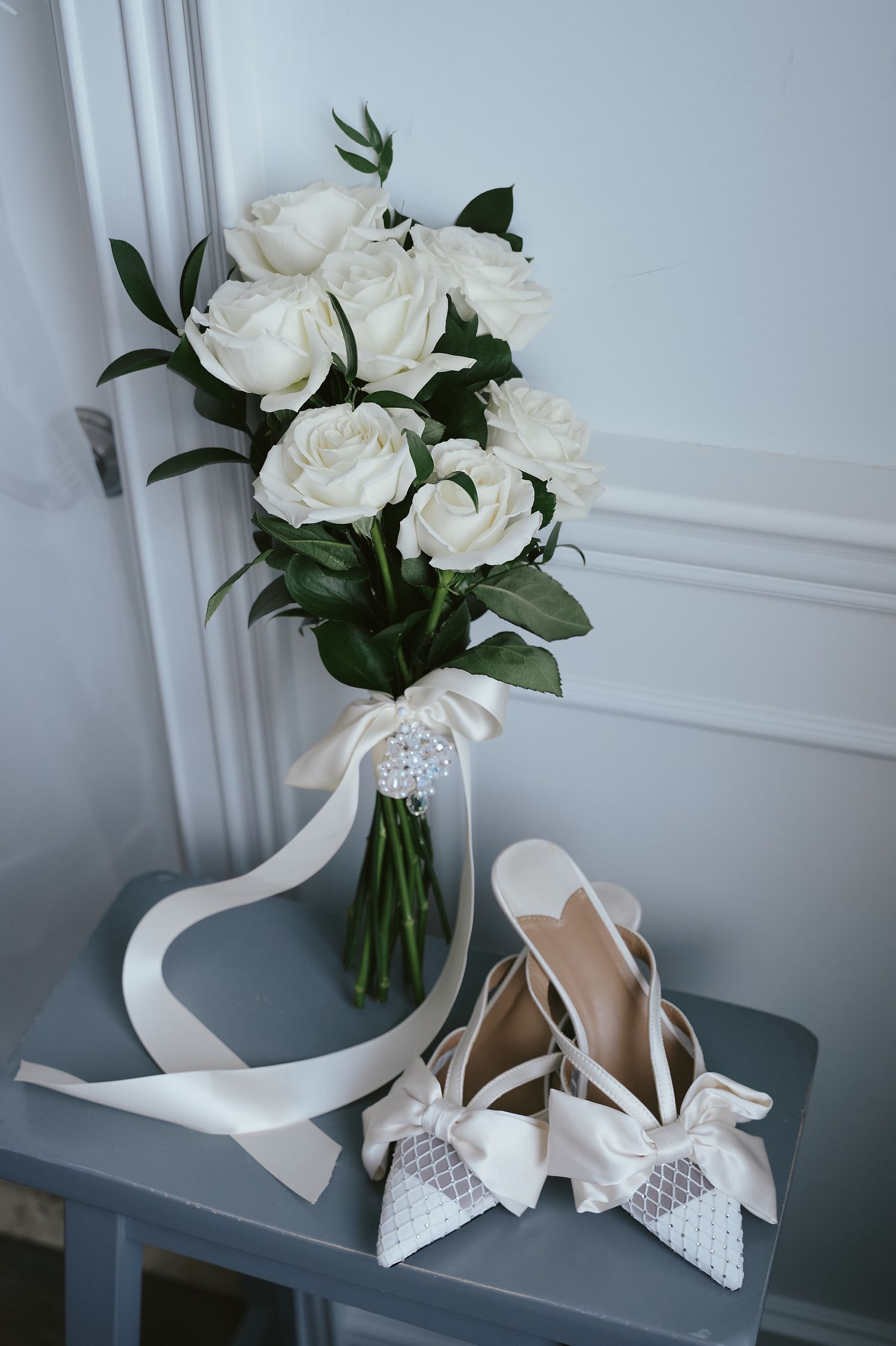 White roses in a vase with greenery alongside elegant bridal shoes with bows and rhinestone details on grey table.