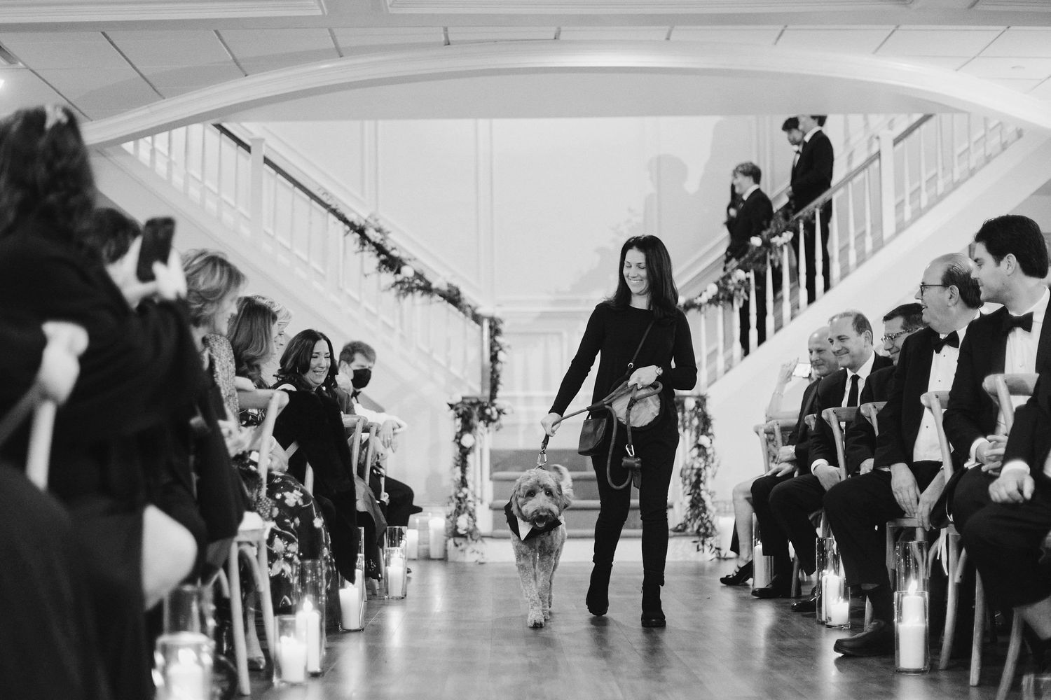 Wedding party attendees lined up in two rows as figure in black dances down center of white hallway.