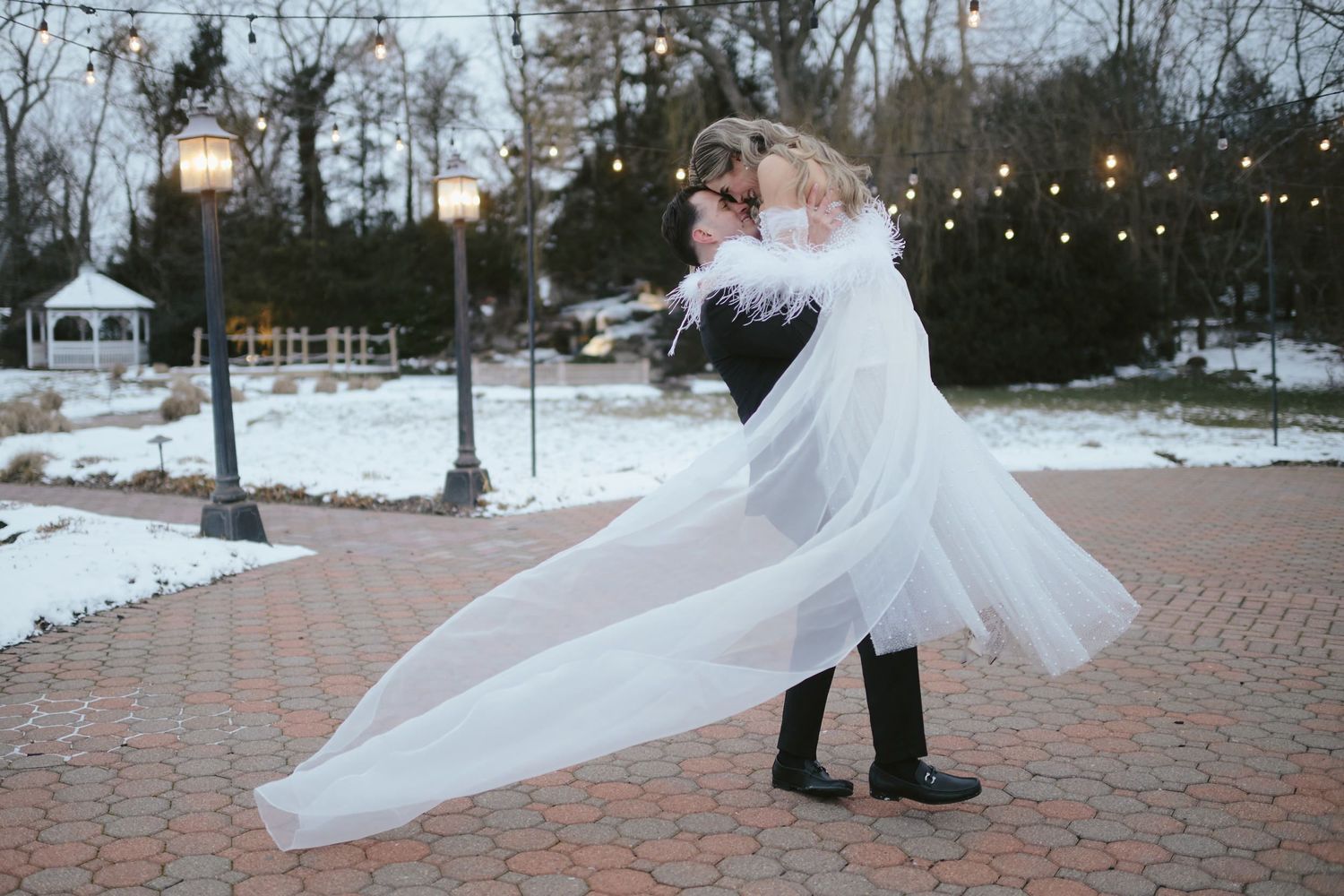 A romantic winter wedding moment captured on snowy brick path with string lights and flowing veil in the evening.