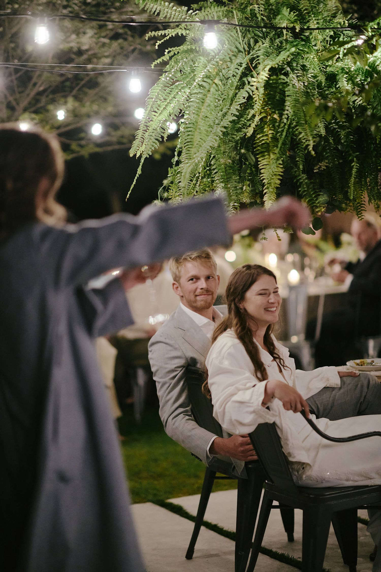 Outdoor evening wedding reception under string lights and fern decorations with guests seated at tables beneath twinkling lights.