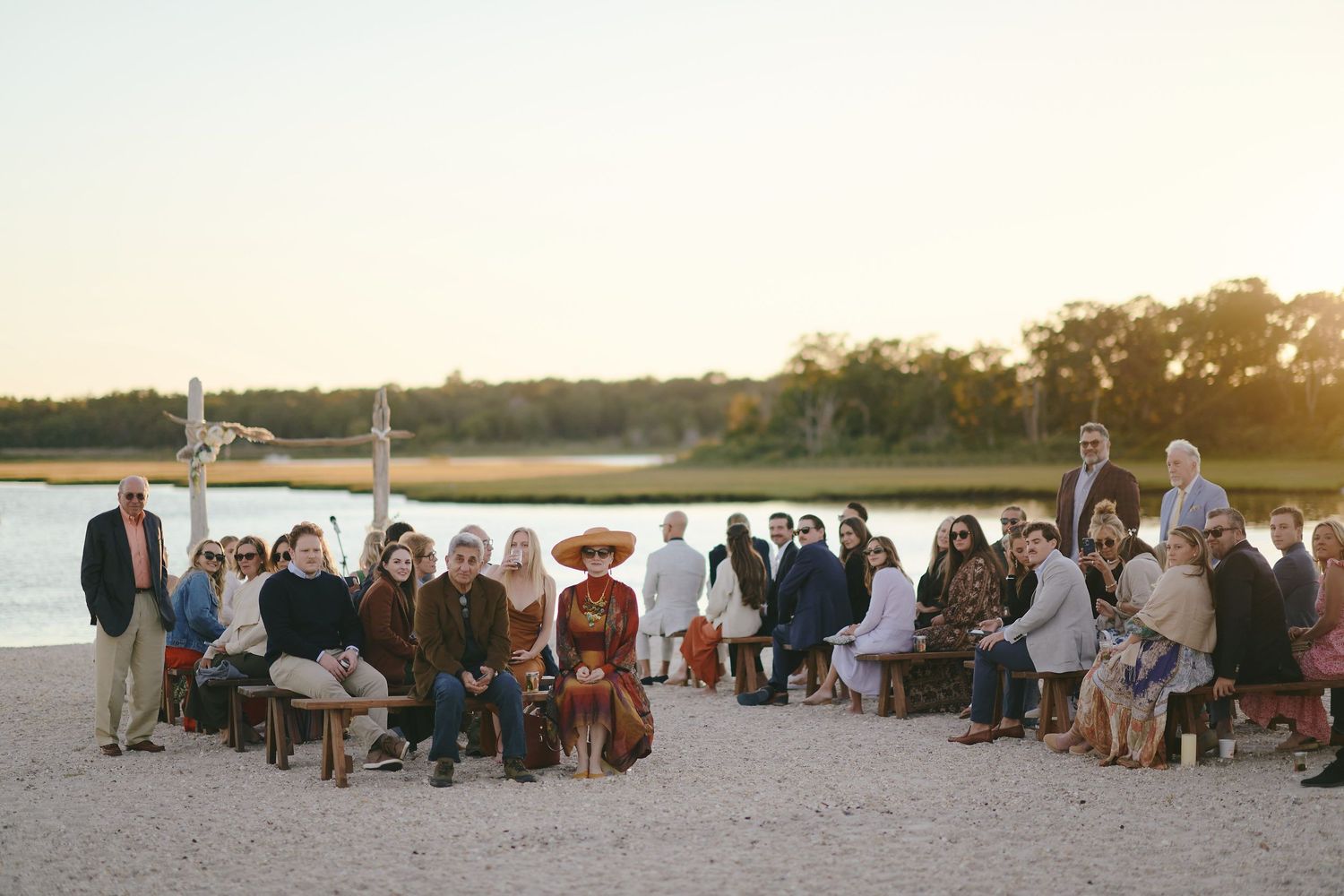 Large group of wedding guests gathered on a beach at sunset near the water and trees.