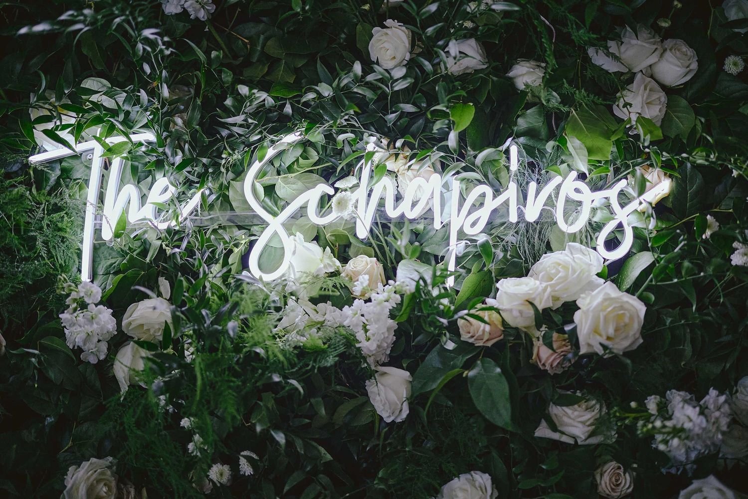 Neon white light sign saying 'The Somewhere' mounted on a green leafy wall with white floral decorations.