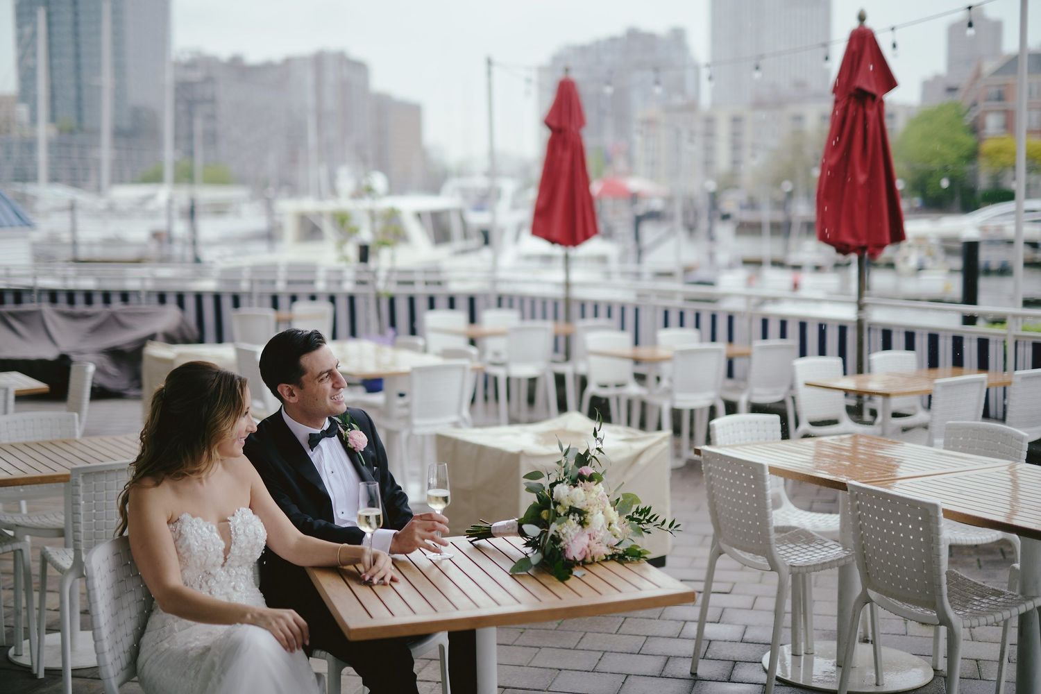 A couple in wedding attire sits at a table on an outdoor patio with red umbrellas and white chairs.