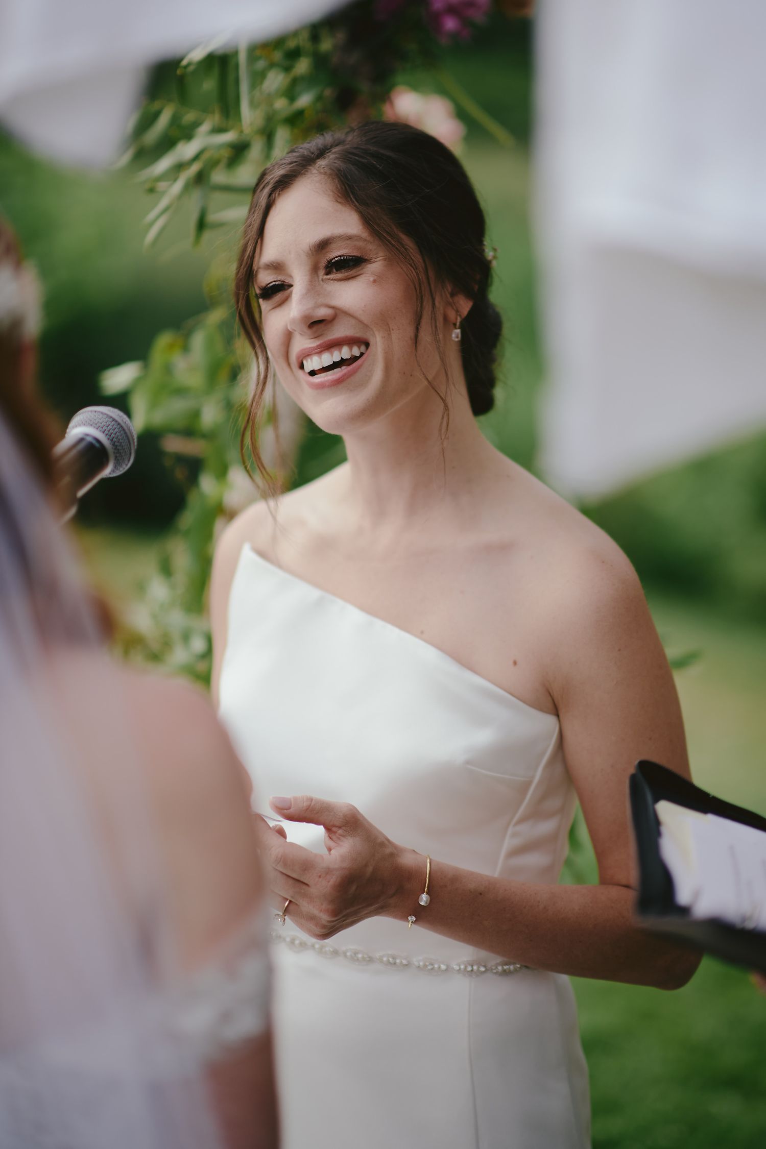 A joyful bride in a strapless white gown laughs while holding papers during an outdoor wedding ceremony.