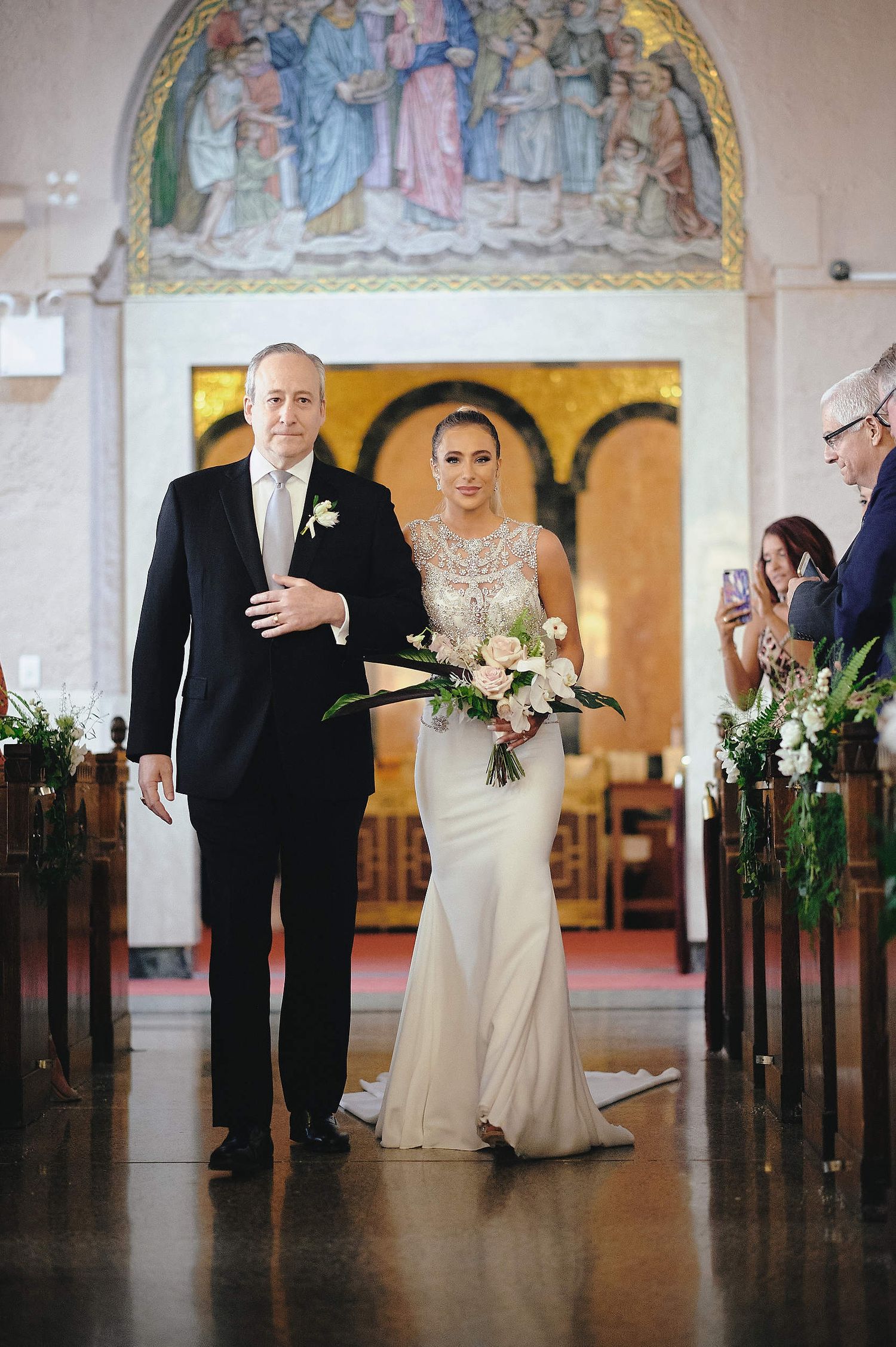 A traditional church wedding ceremony with a beautiful mural above, decorated pews with greenery lining the aisle.