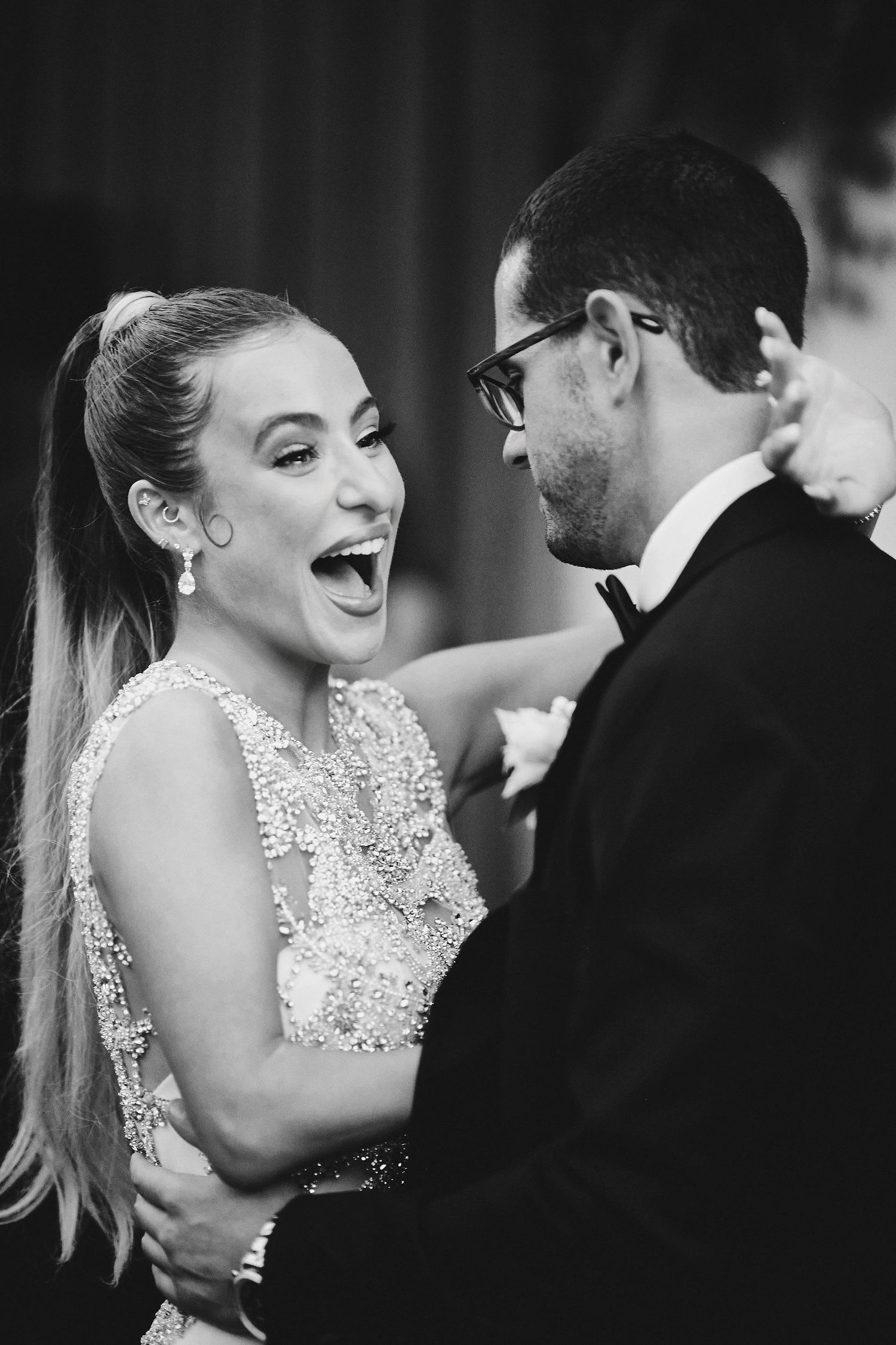 Black and white photo of a couple sharing joyful laughter at their wedding, bride in beaded dress and groom in glasses.