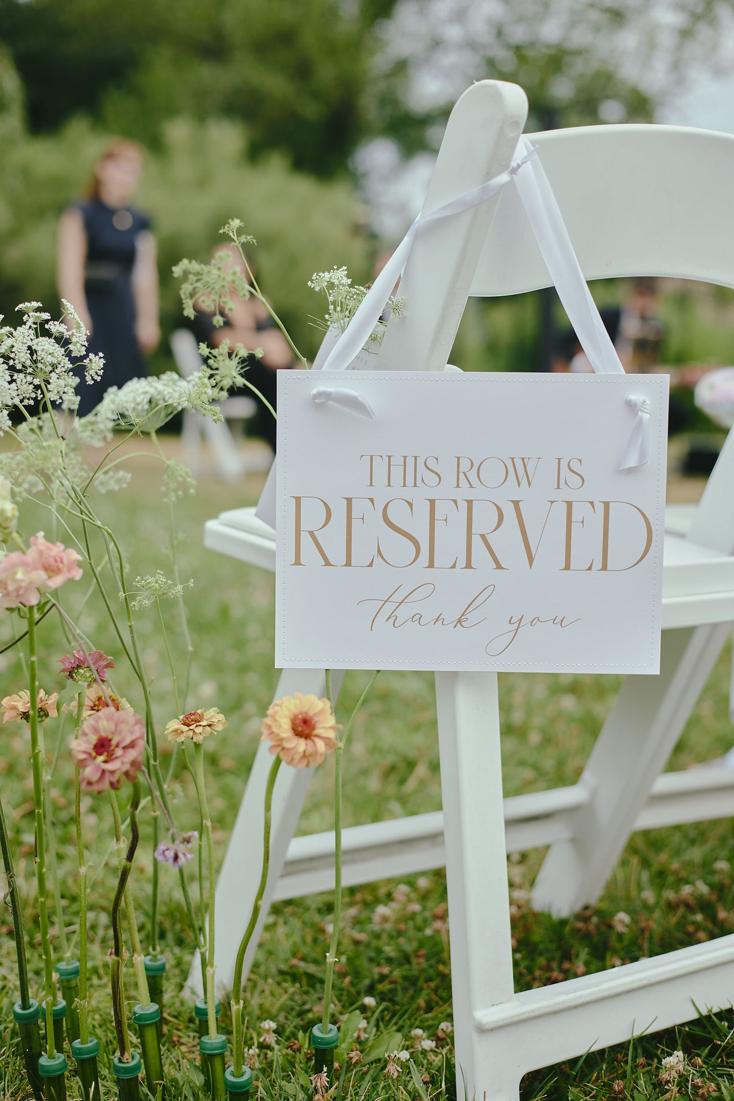 A white 'Reserved' sign hangs on a folding chair at an outdoor wedding ceremony surrounded by wildflowers and greenery.