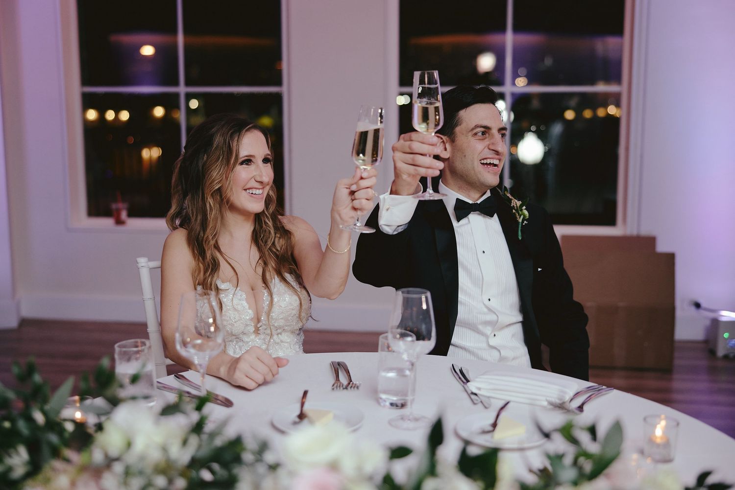 Wedding couple raises champagne glasses while seated at their decorated reception table with candlelight and flowers.