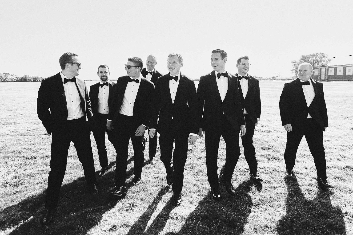 A group of people in formal black tuxedos standing together casting long shadows in black and white photography.