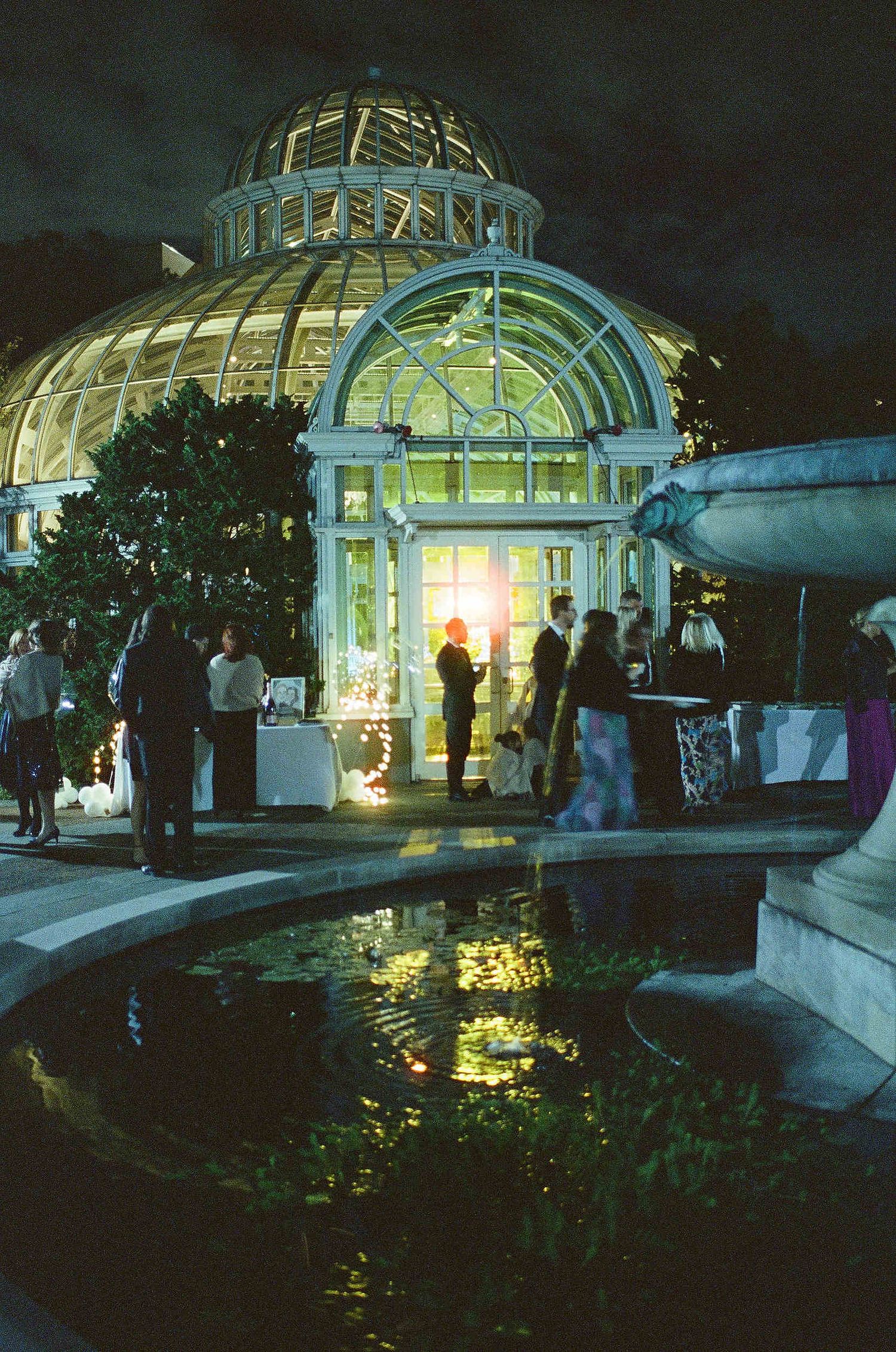Illuminated glass conservatory dome at night with crowd gathered by reflecting pool at botanical gardens or park.