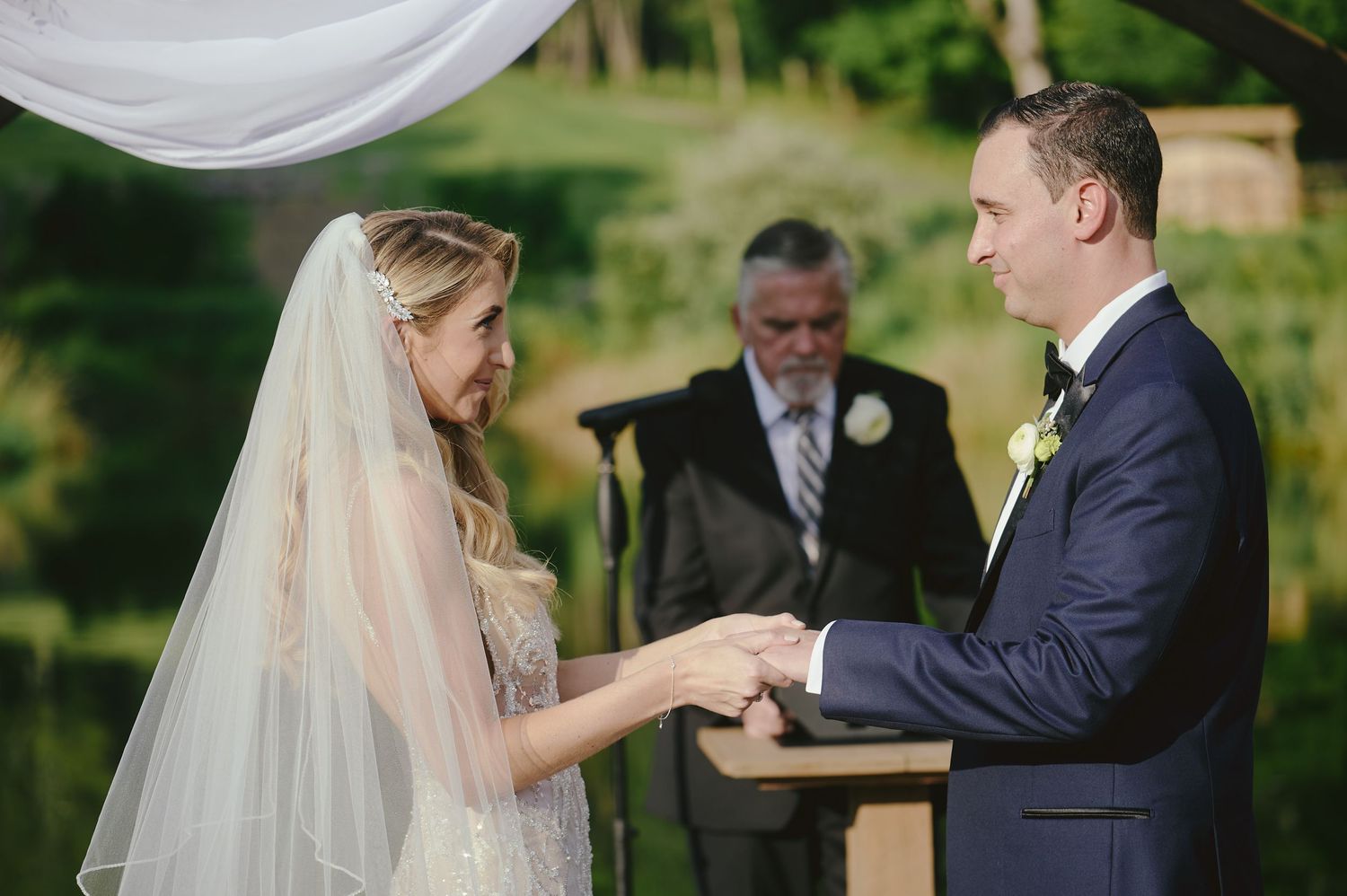 A couple exchanges rings during an outdoor wedding ceremony under a white canopy with a wedding officiant present.