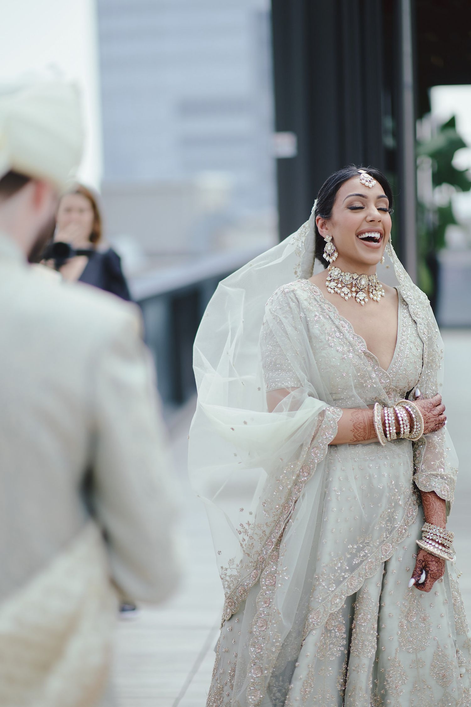 A bride in an elegant white embroidered lehenga and flowing veil reacts joyfully during a traditional Indian wedding ceremony.