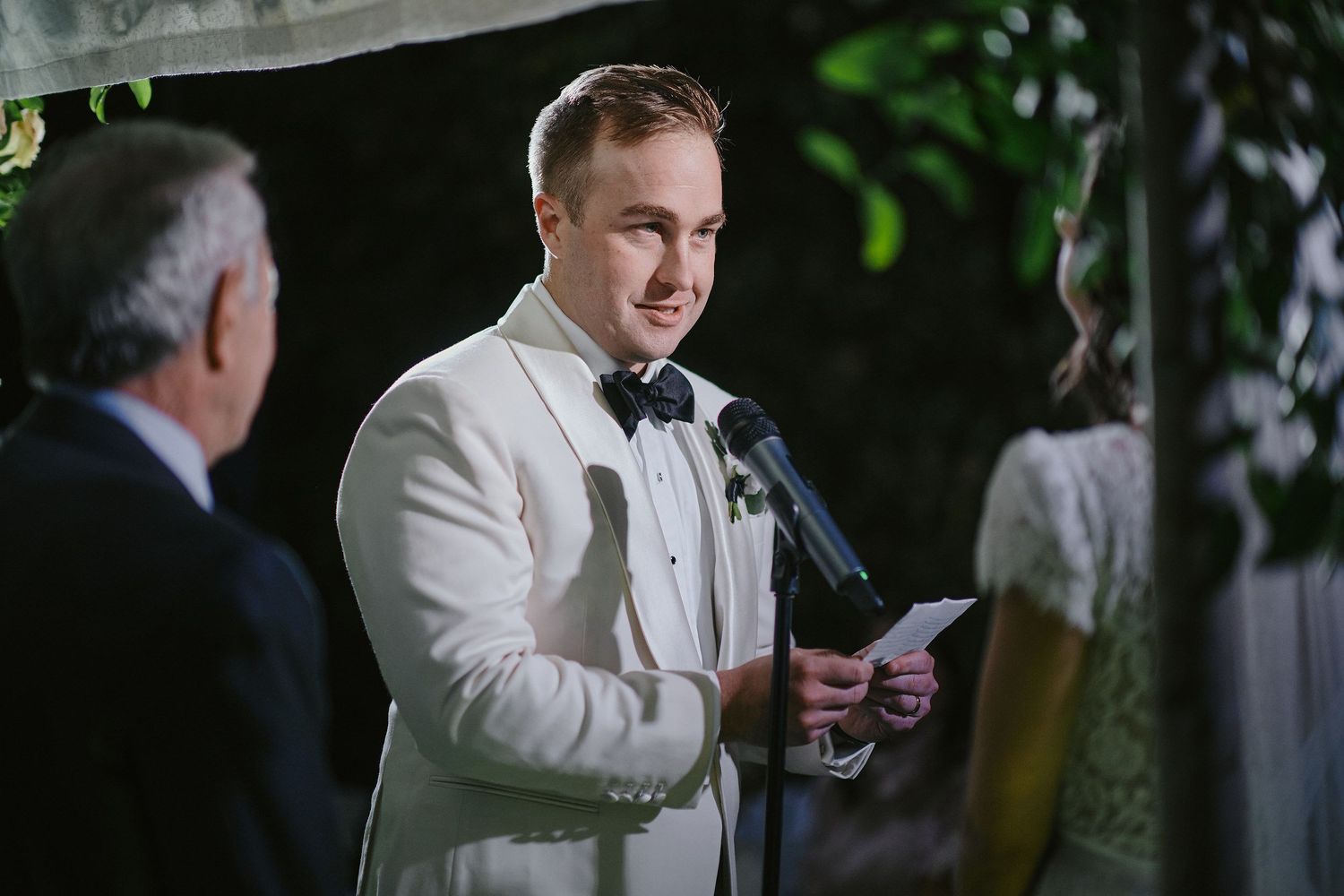 A person in a white tuxedo jacket and bow tie reads from a paper while speaking into a microphone during an event.