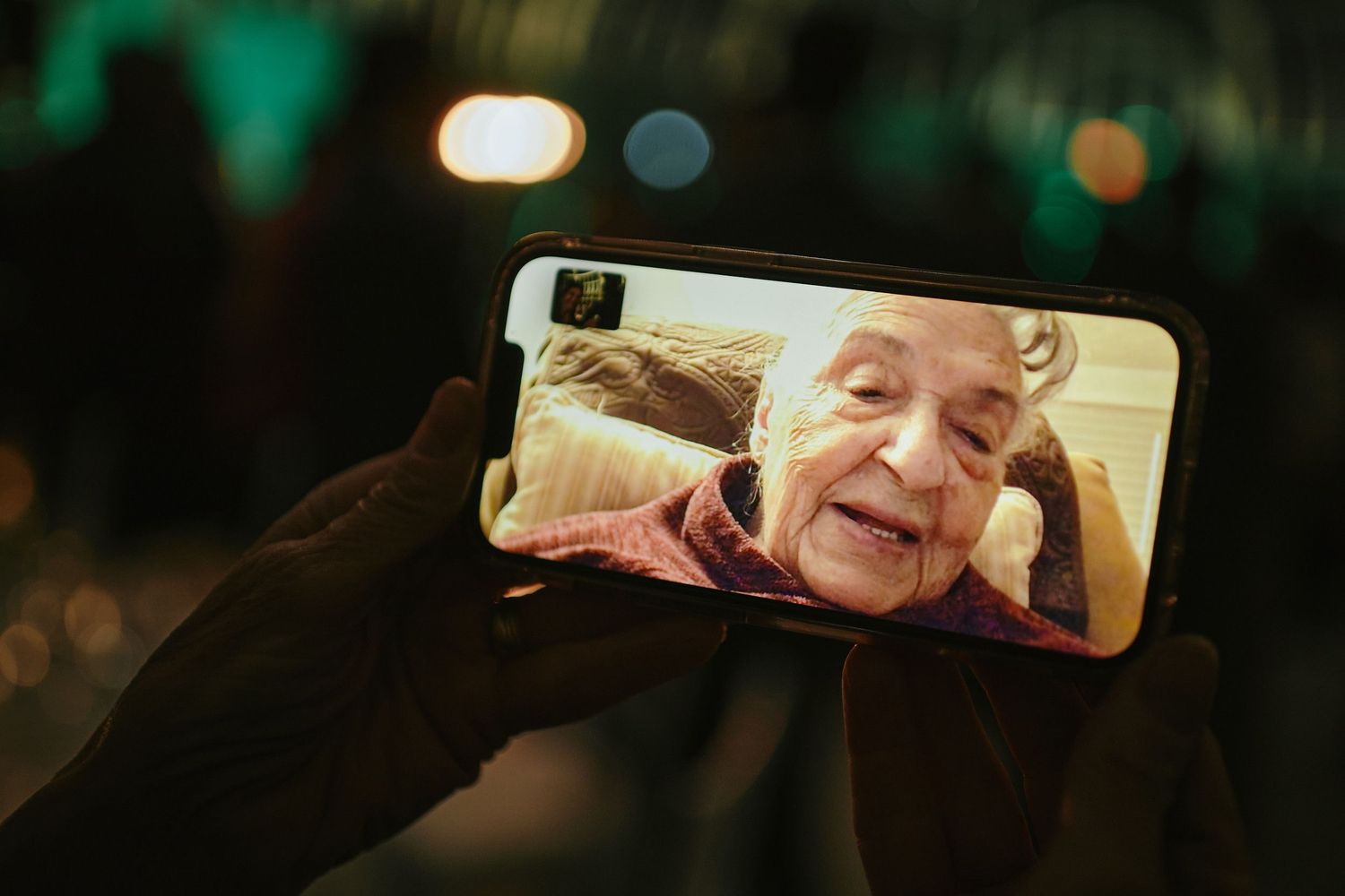 Someone holds a smartphone displaying a video call on a dark background with blurred lights.