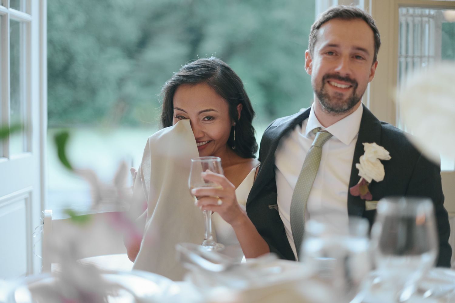 Bride and groom smiling during wedding reception toast, bride holding champagne glass and napkin to face.