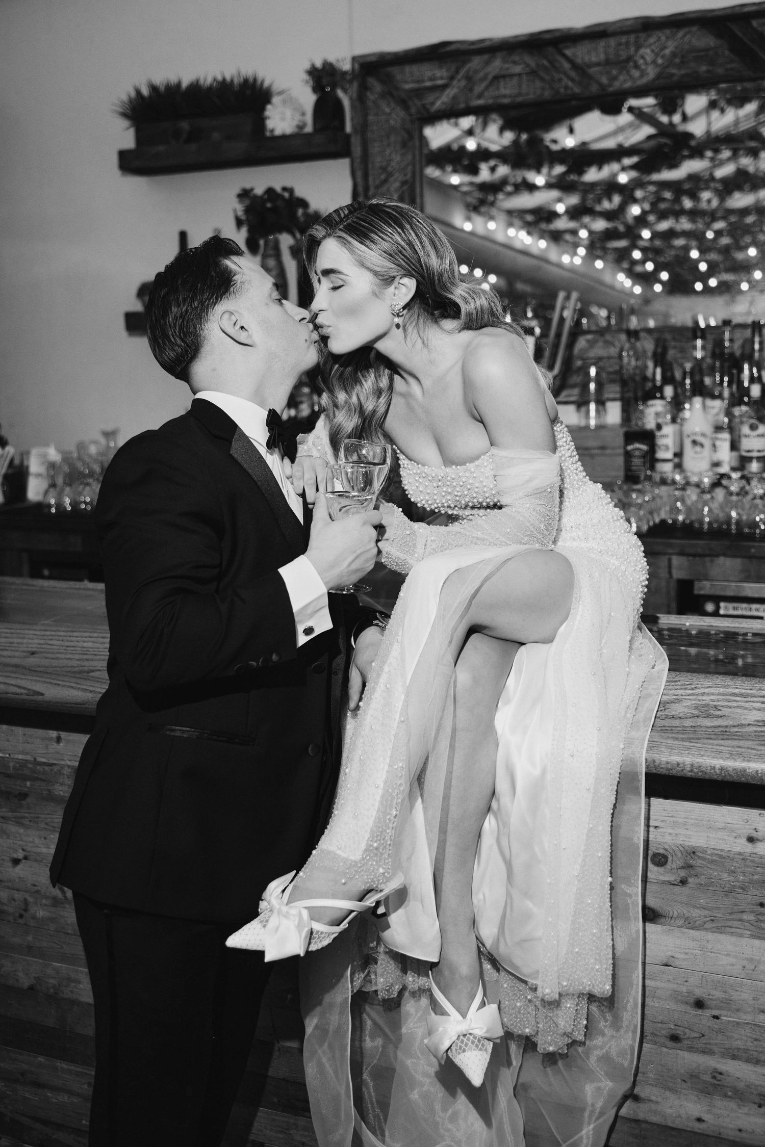Black and white photo of a bride and groom kissing while holding champagne glasses at their wedding reception.