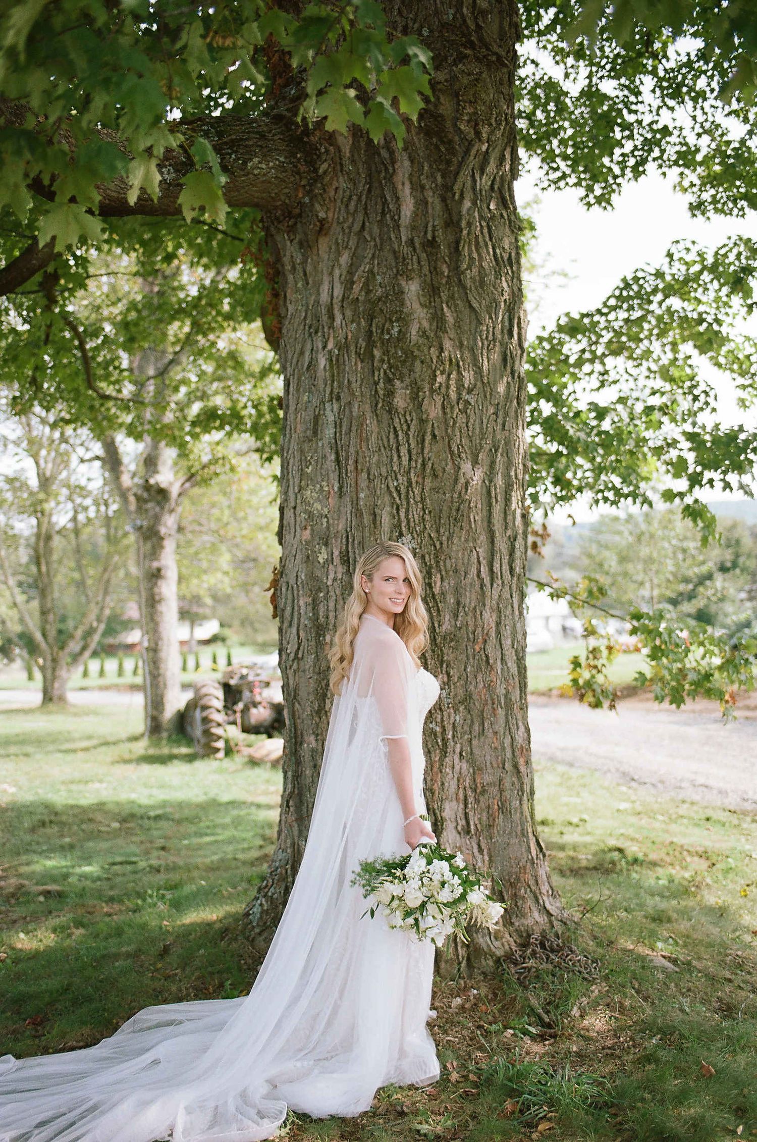 A bride in a flowing white gown with a long train stands beside a large tree trunk holding a floral bouquet outdoors.
