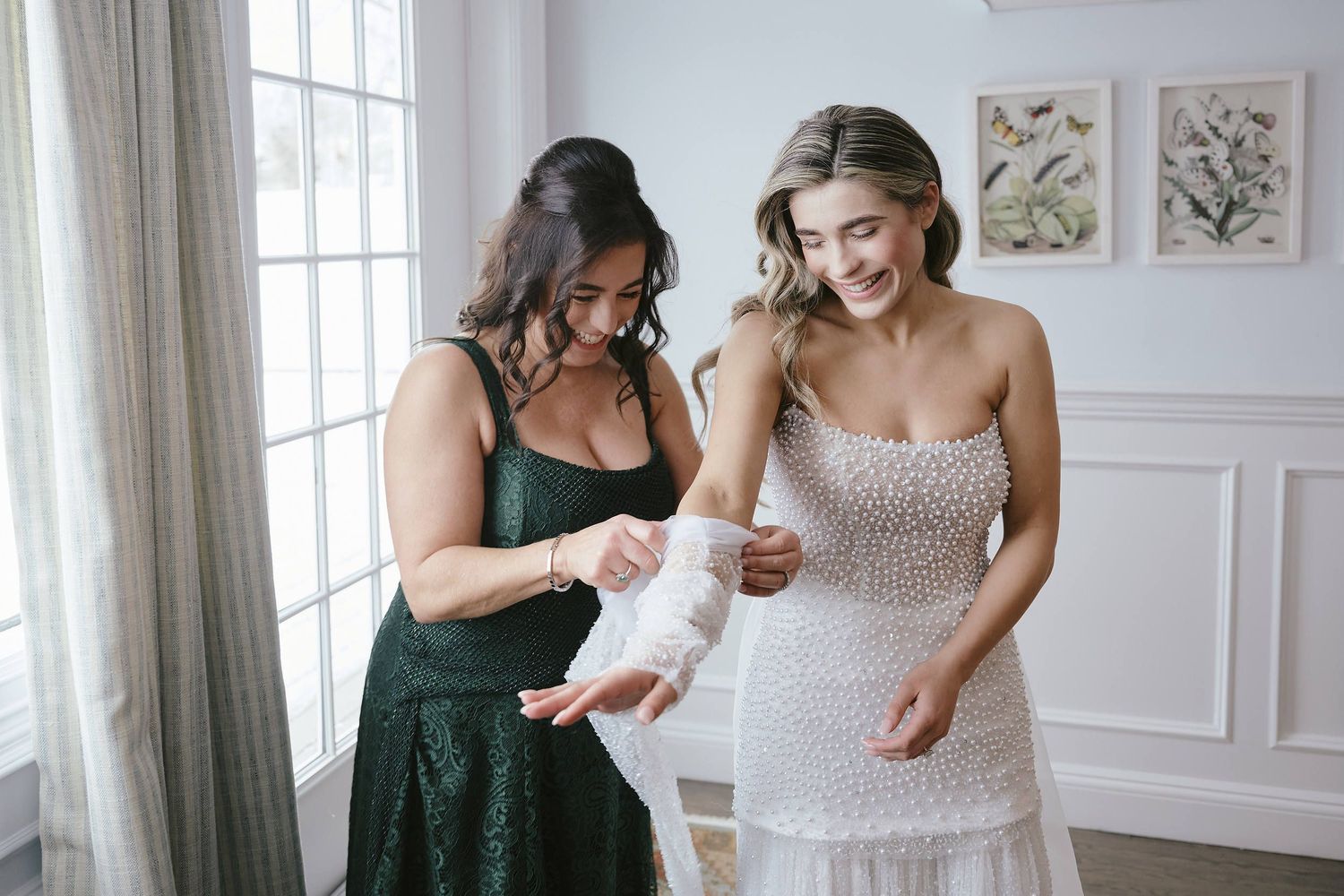 Two women in elegant dresses smile while looking at a document together in a bright room with botanical prints.