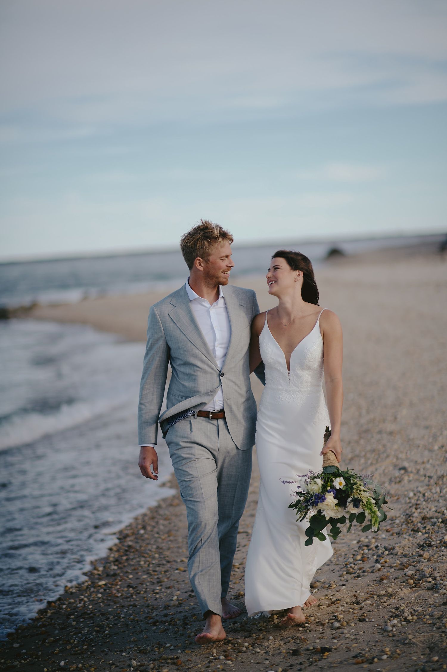 Newlywed couple walking barefoot on a scenic beach at sunset with waves and a bouquet of white flowers.
