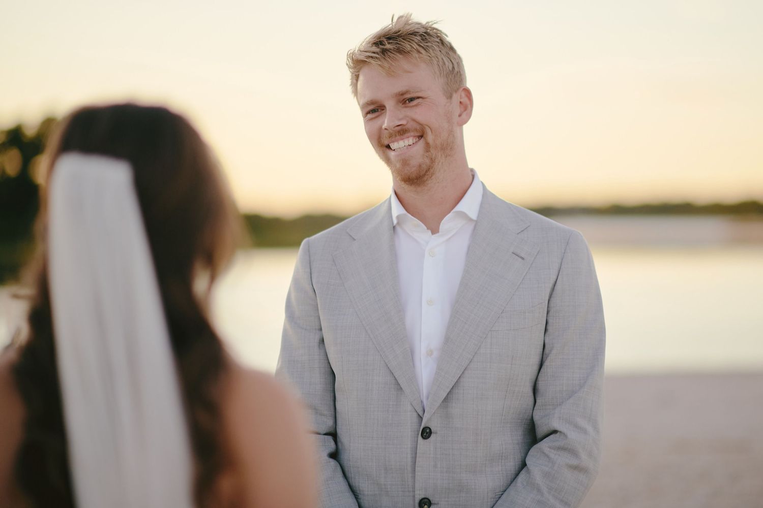 Close up wedding ceremony scene of groom in light gray suit smiling during outdoor sunset ceremony by water.