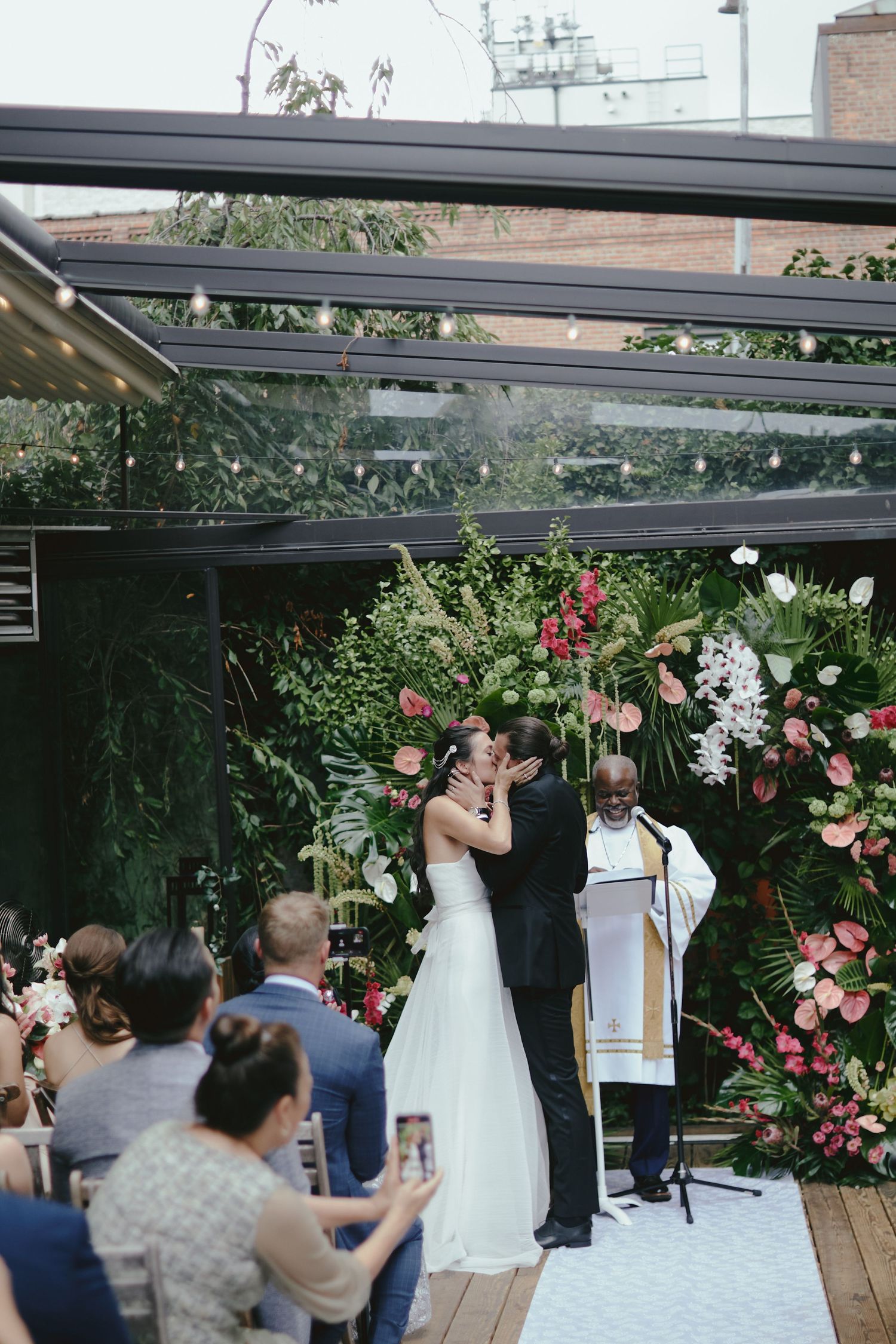 Newlyweds share their first kiss at wedding ceremony under covered patio with pink floral arch and guests watching.