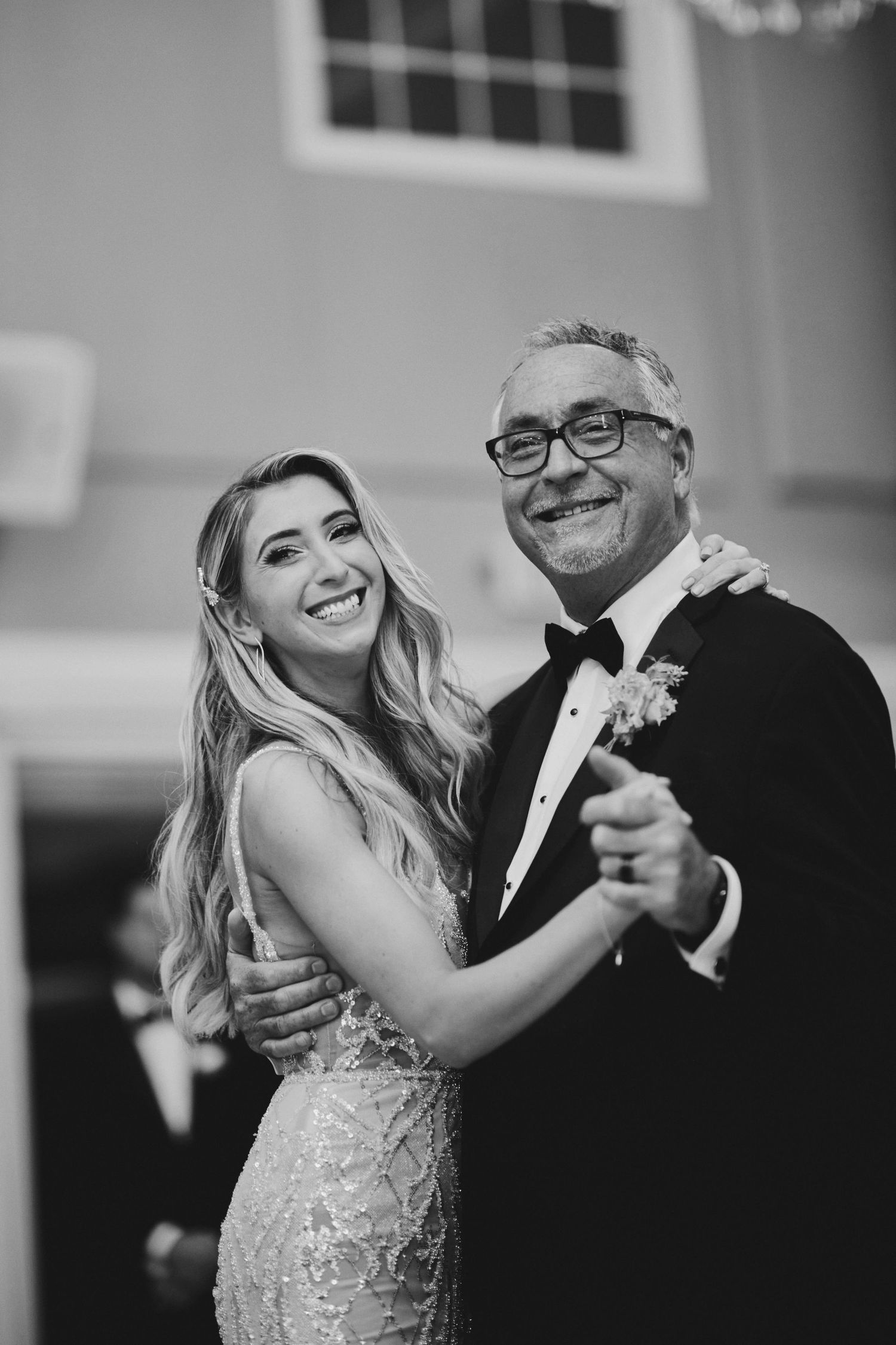 A bride in a lace dress and older companion share a joyful dance together at a reception in black and white.