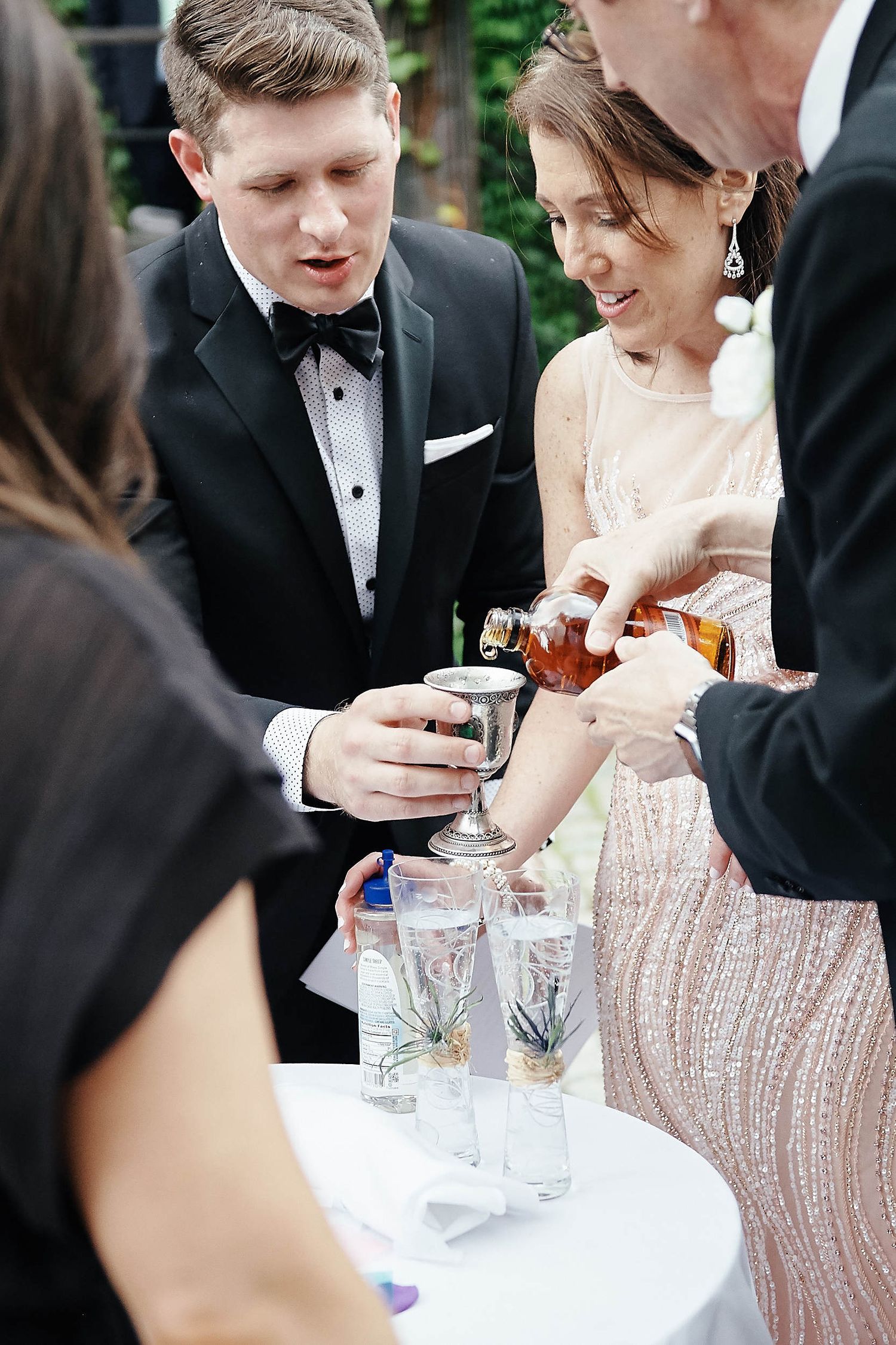 Guests pouring drinks at an outdoor wedding reception with crystal glassware on white table.