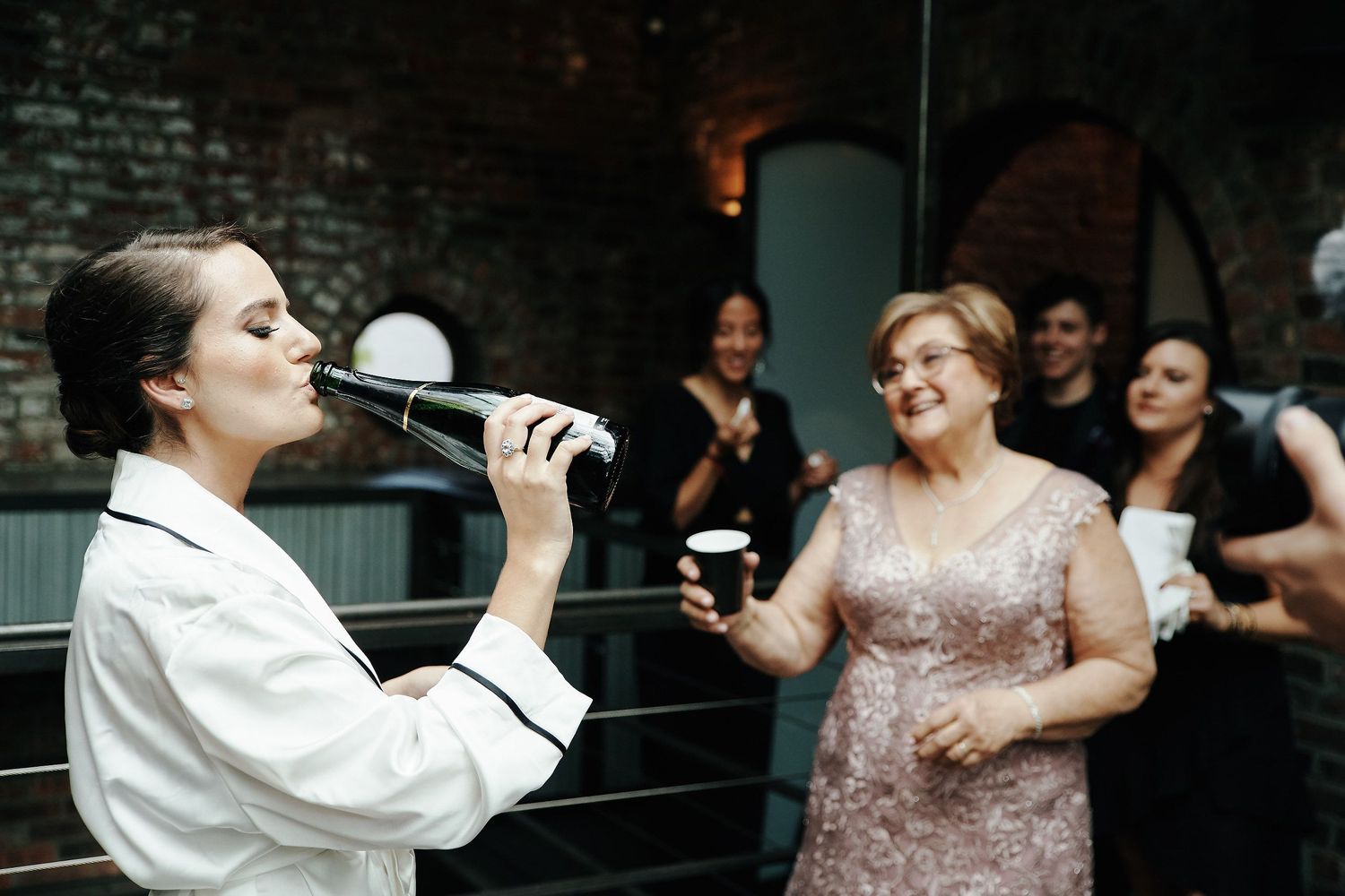 Two people interacting joyfully at an indoor event, one in white holding a microphone and one in a sparkly rose-colored dress.