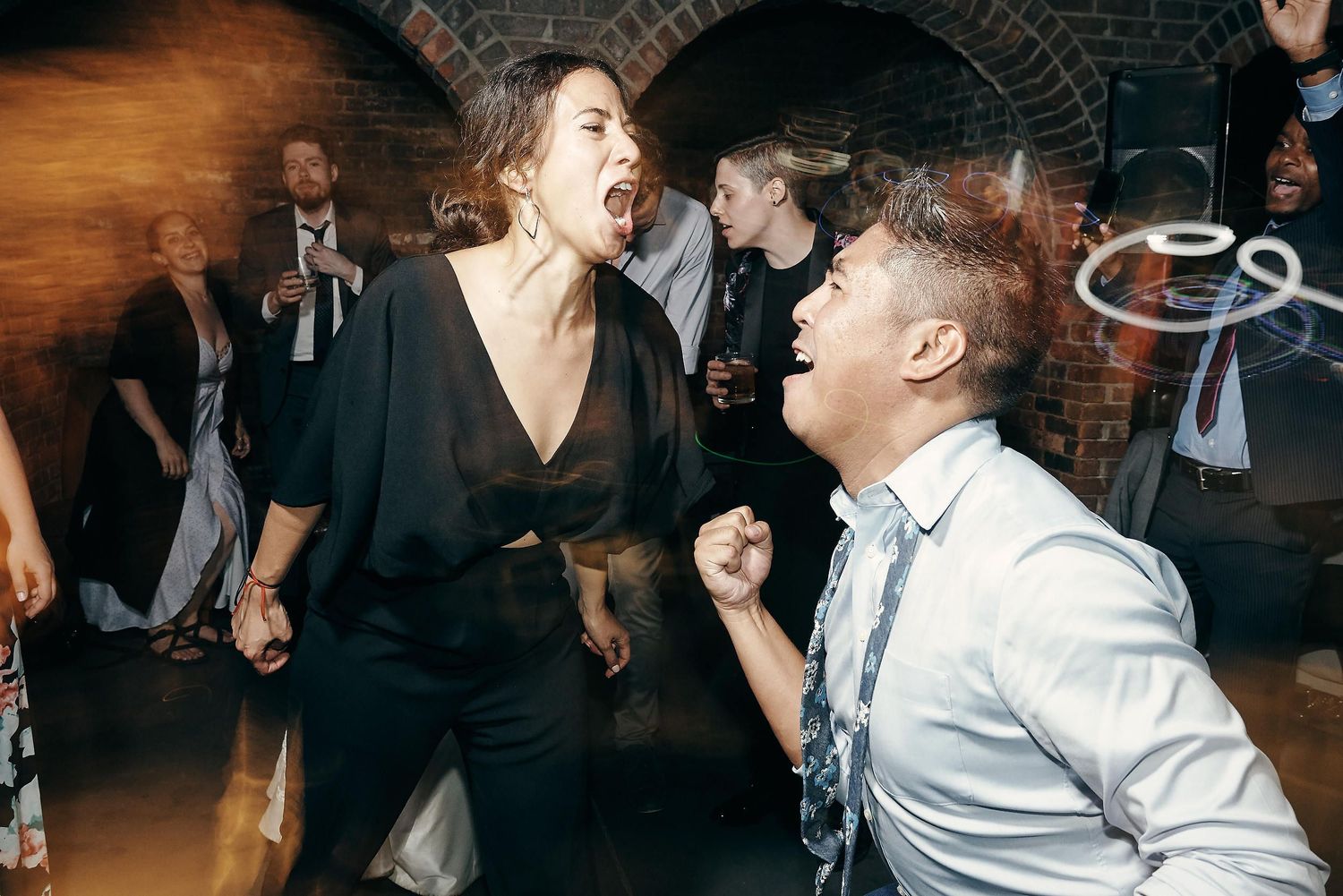 Wedding guests dancing and celebrating energetically at a dimly lit reception venue with brick walls.
