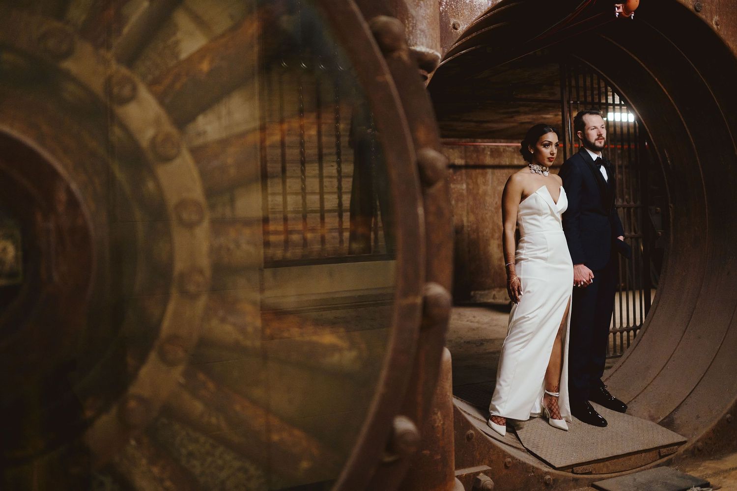 A couple in formal wedding attire poses in an ornate vault-like doorway with moody lighting.