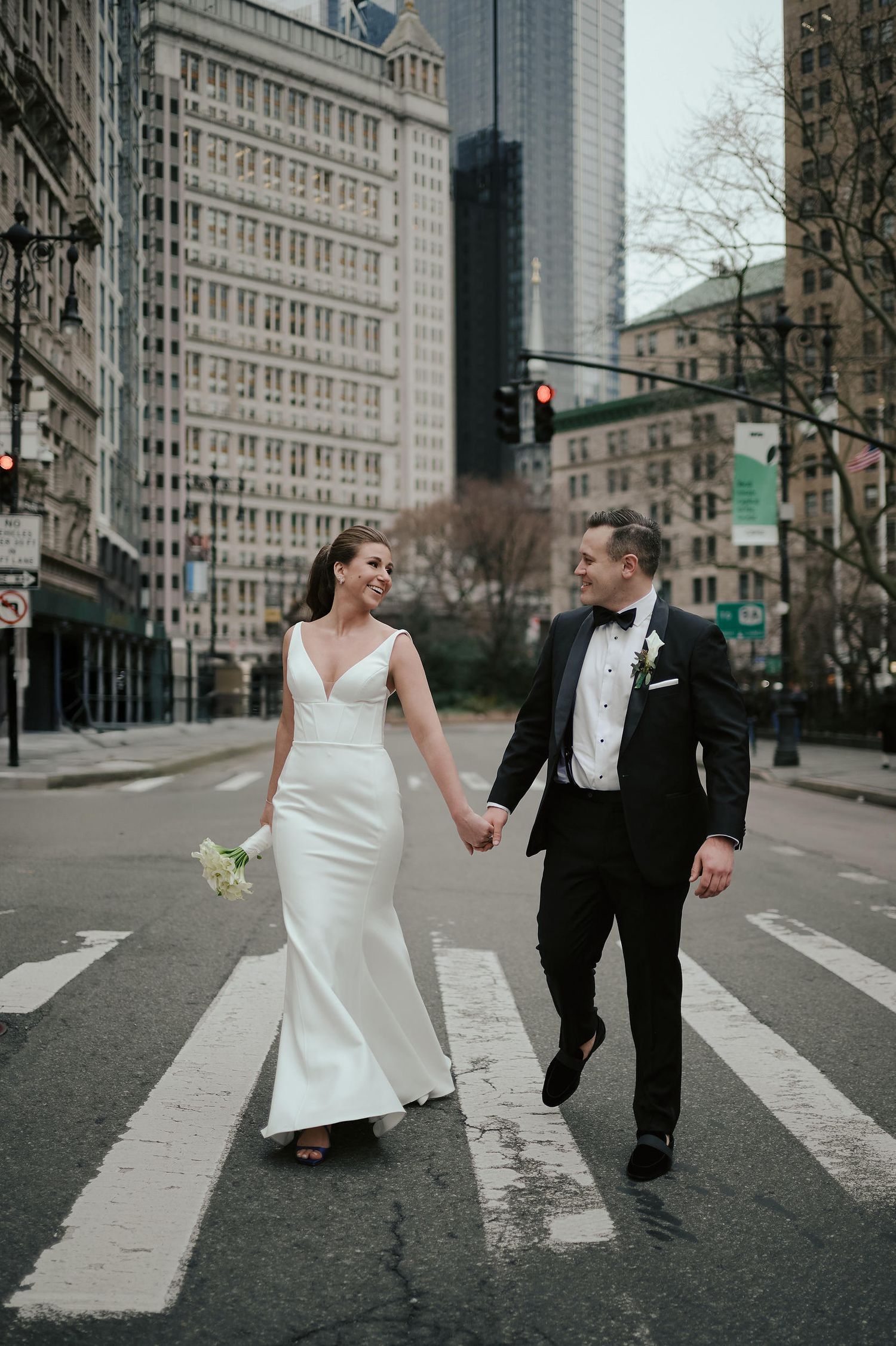 A couple in wedding attire walks across a crosswalk in downtown with tall buildings in the background.