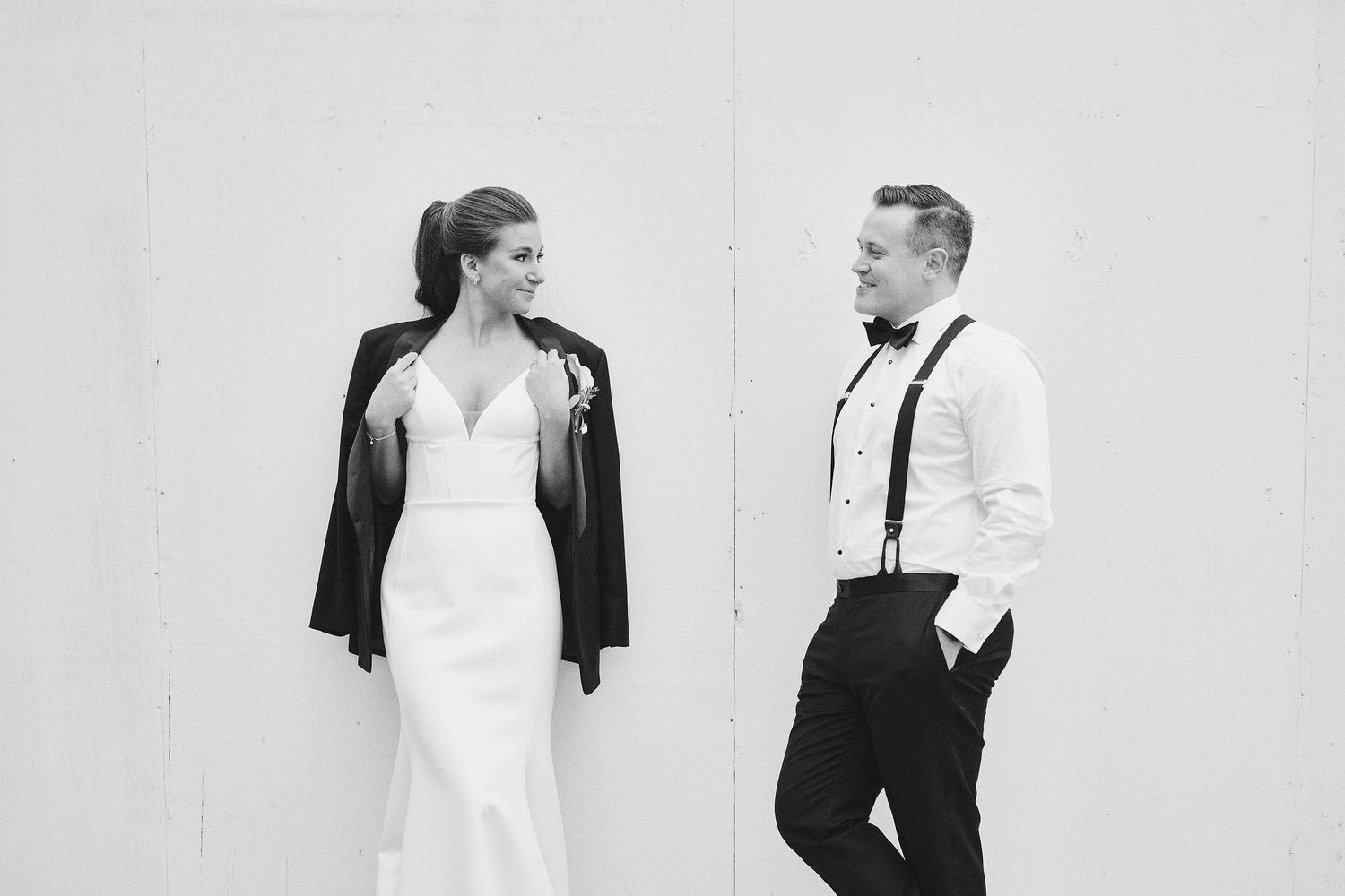 A bride in a fitted white dress and black jacket poses with groom in white shirt and suspenders against white wall.