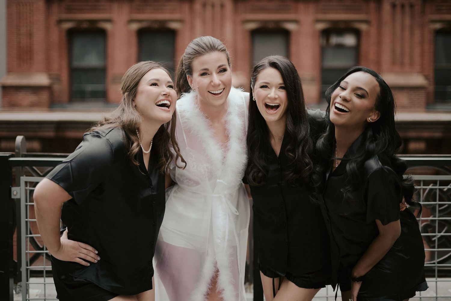 Bride in white dress shares joyful laughter with bridesmaids in black dresses outside a brick building.