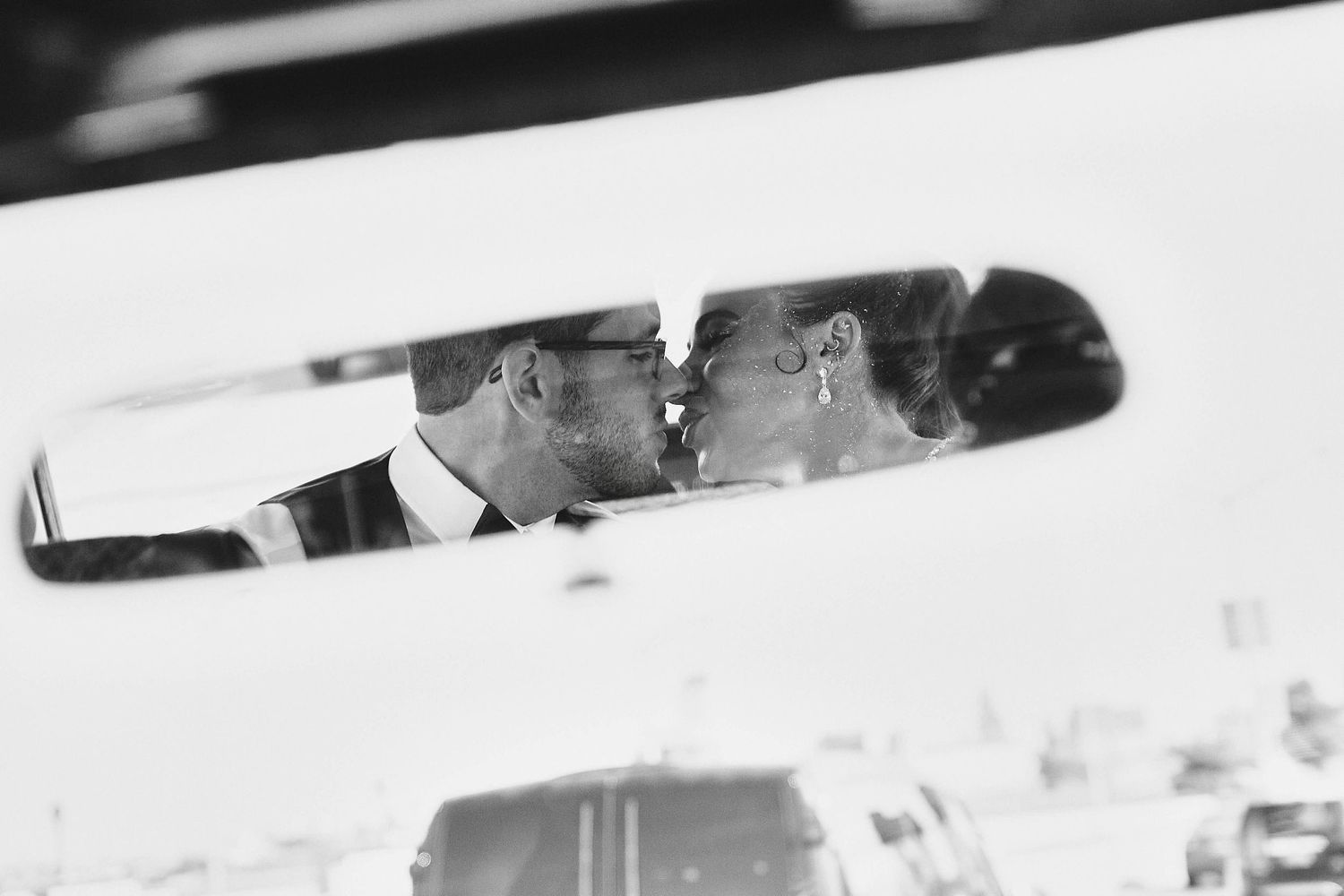 A romantic couple kisses while their reflection appears in the rearview mirror of a car.