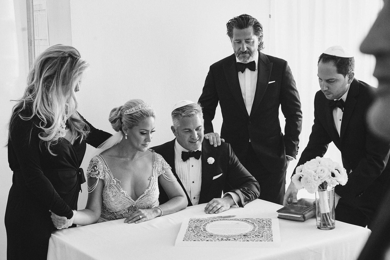 Elegant black and white photo of wedding party gathered around table signing documents during formal celebration.