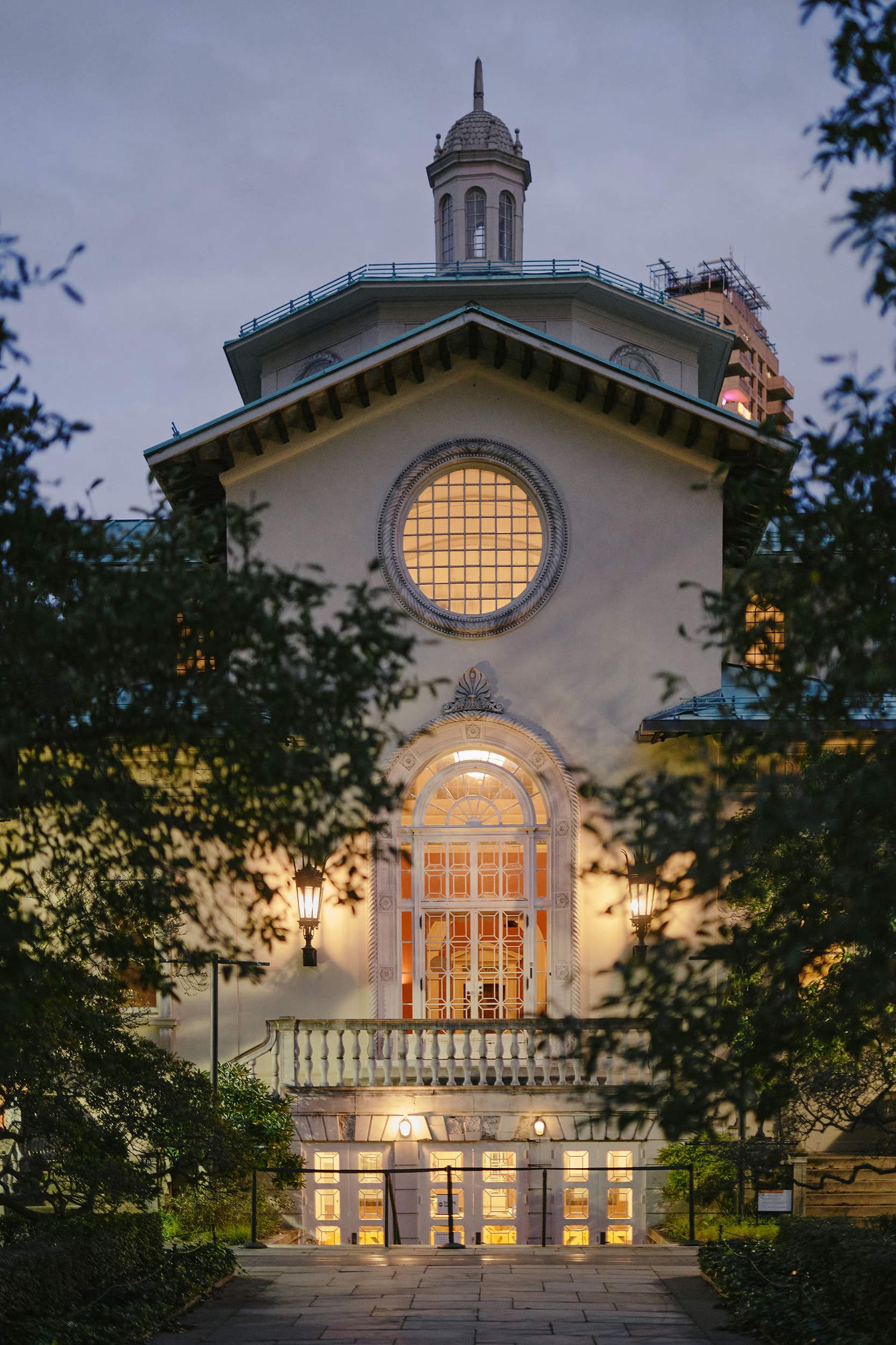 Historic church facade with illuminated circular window and arched entrance glows warmly at dusk against a cloudy sky.