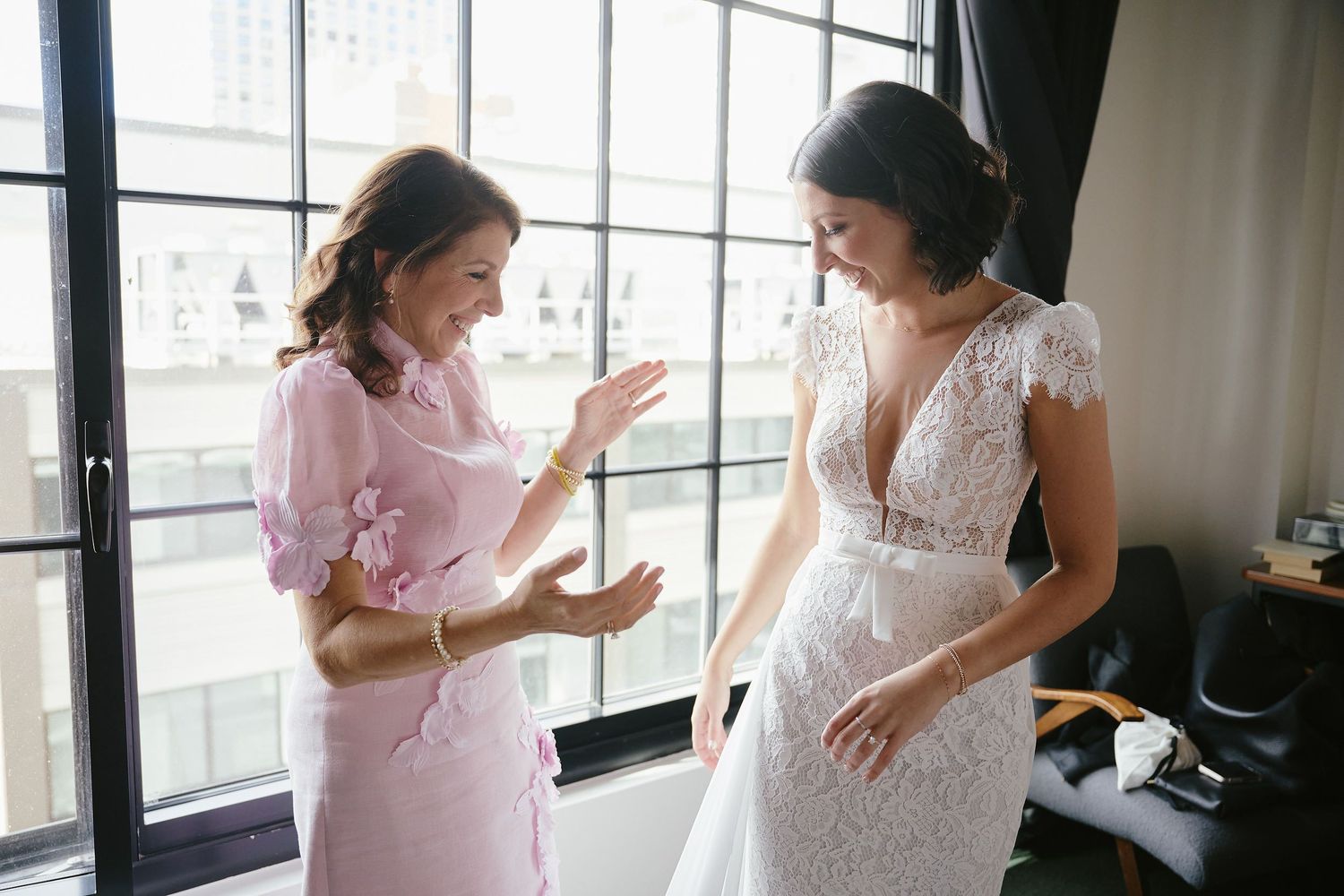 Two people share an emotional moment as they prepare for a wedding, one wearing a pink dress and one in a white lace gown.