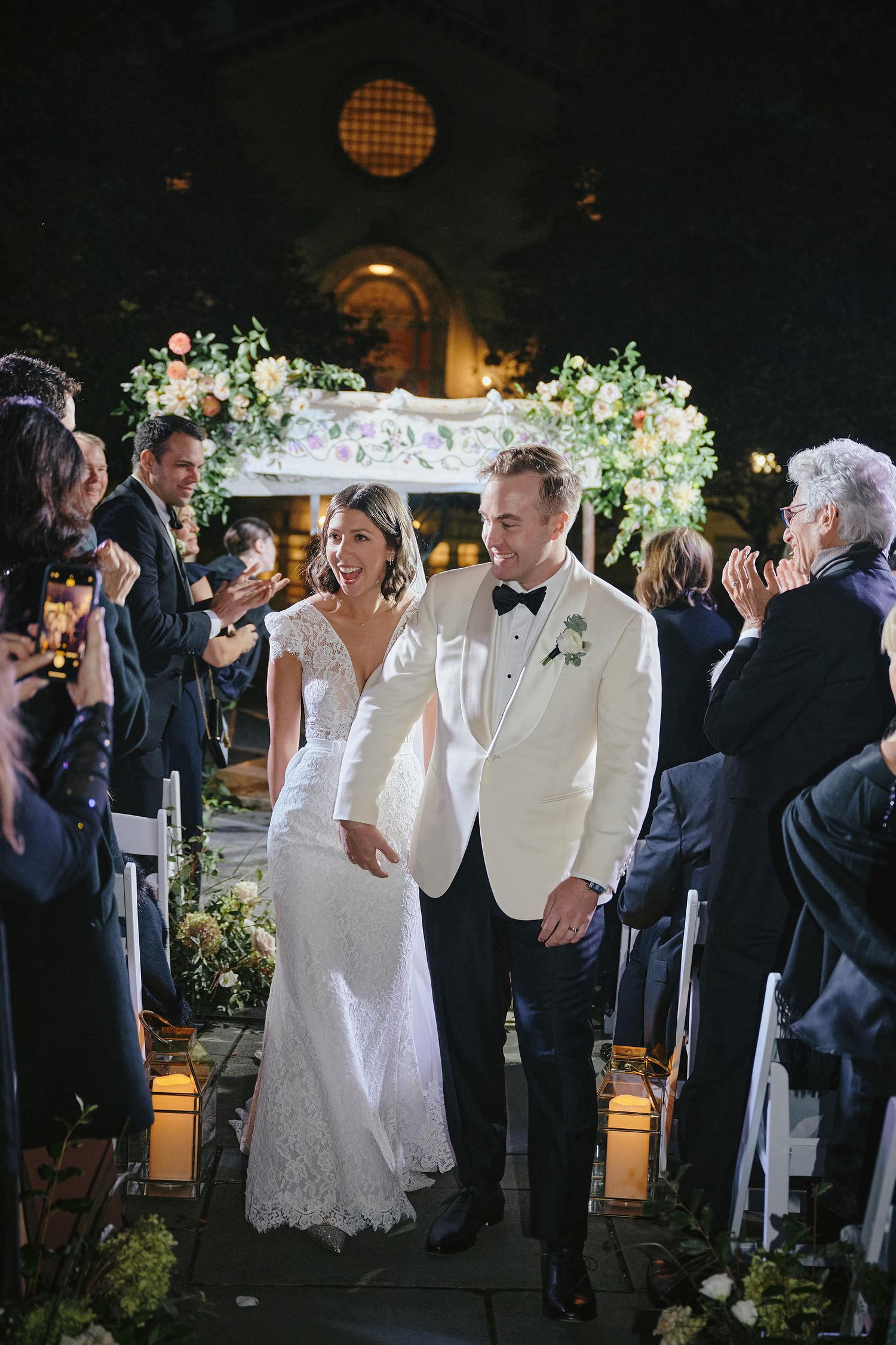 A romantic wedding ceremony in a dimly lit venue decorated with elegant floral arrangements and lanterns along the aisle.