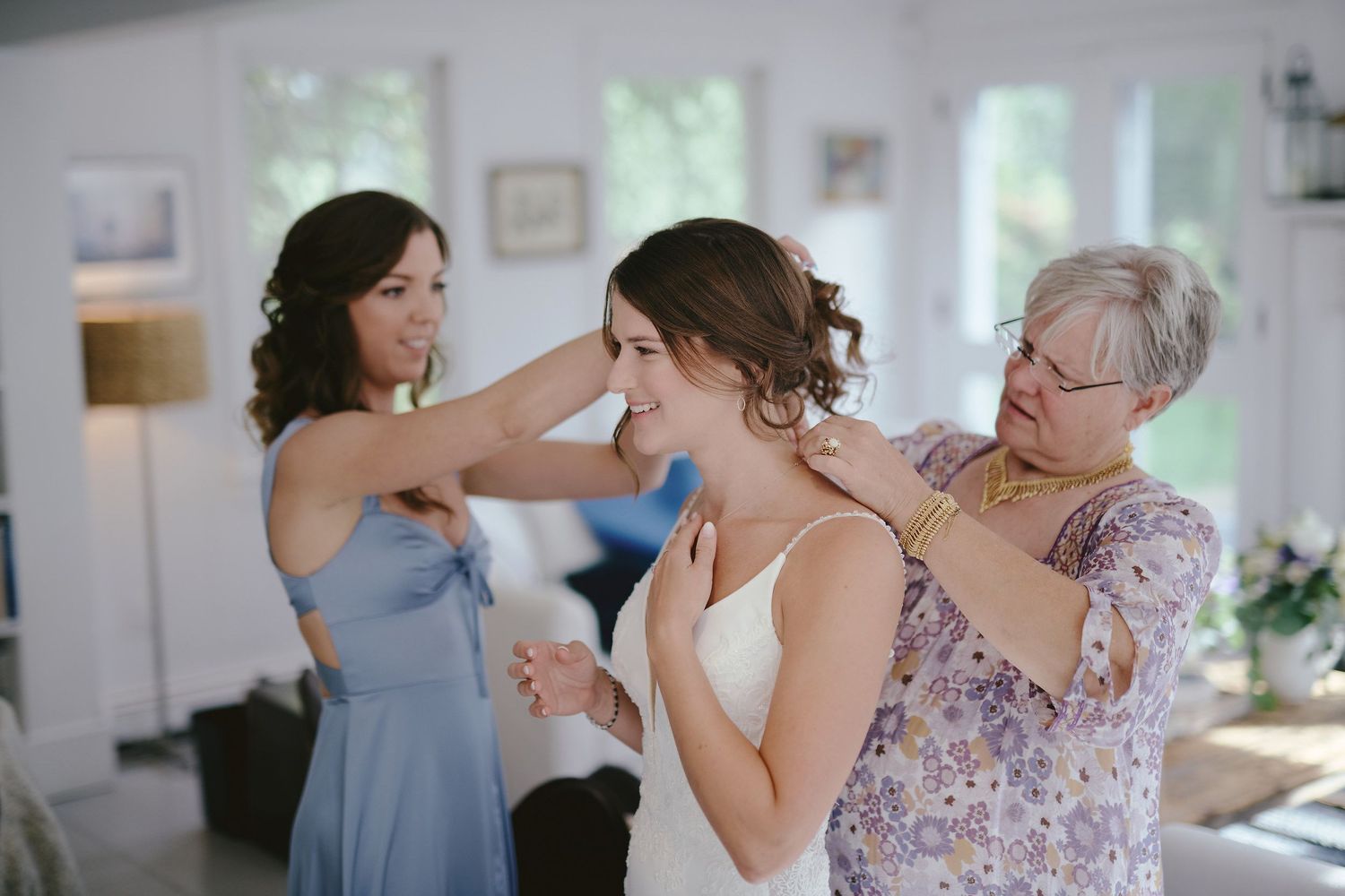 A bride getting ready on her wedding day with two helpers assisting with final preparations in a bright room.
