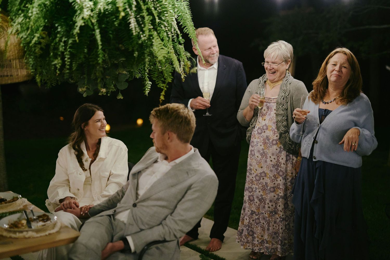 Small group of people gathered at an outdoor evening social event with hanging plants in the background.