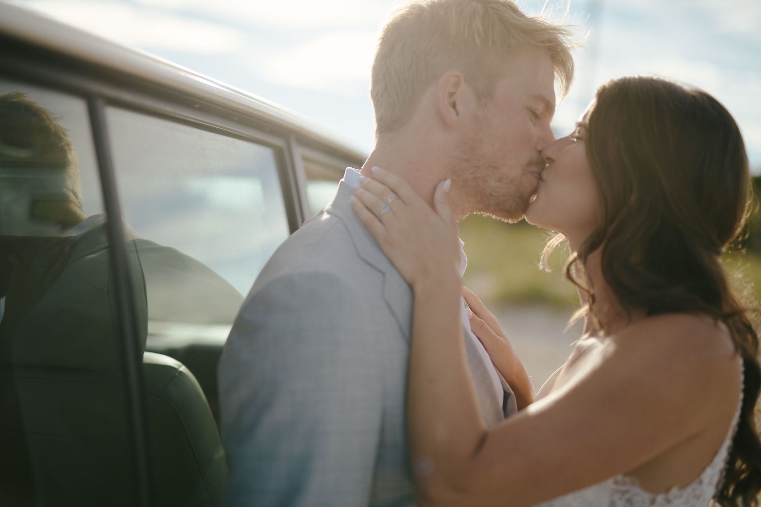 A romantic kiss between a couple in formal attire next to a car on a sunny day.