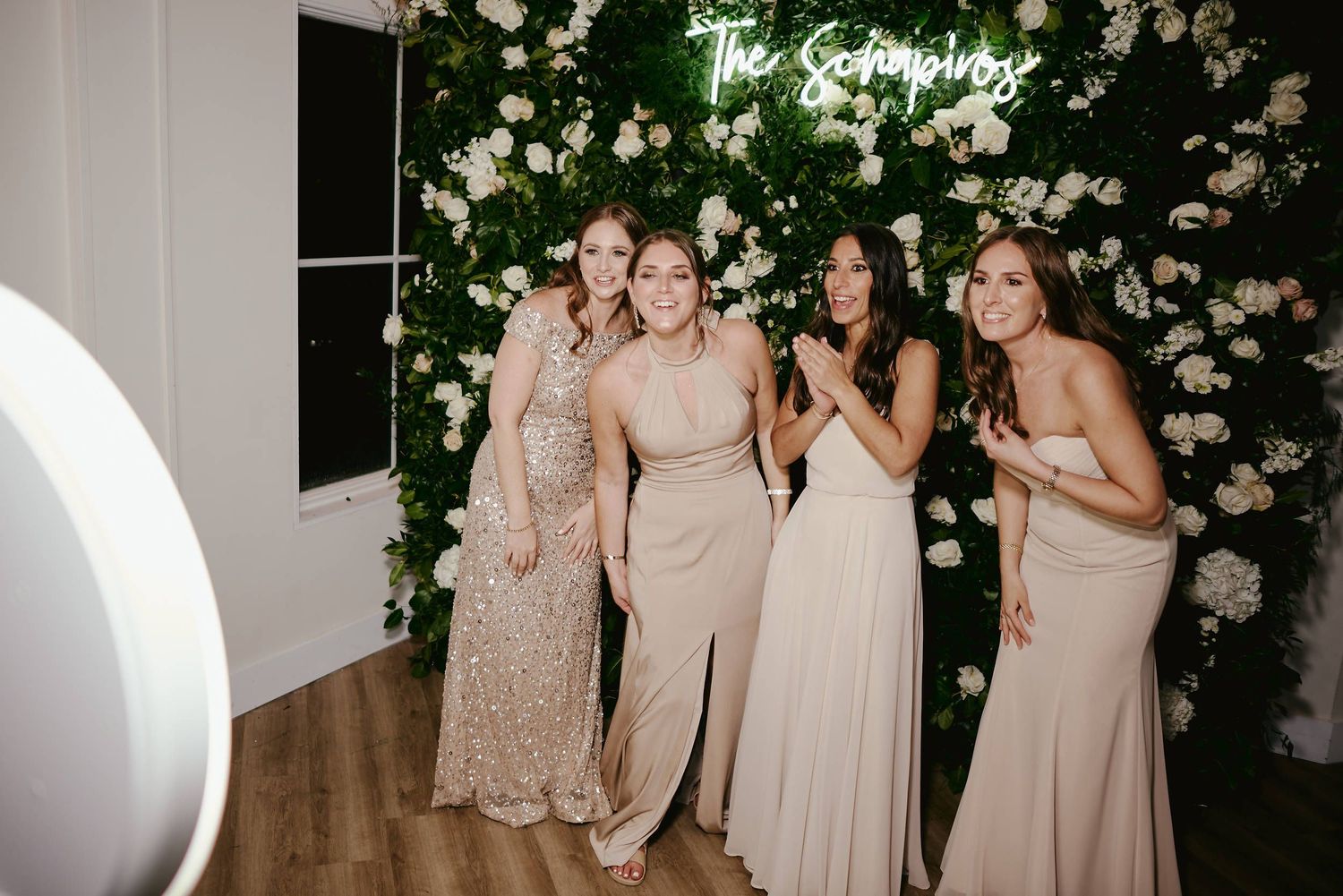 A group in elegant neutral-toned gowns pose together against a white floral backdrop at an evening event.