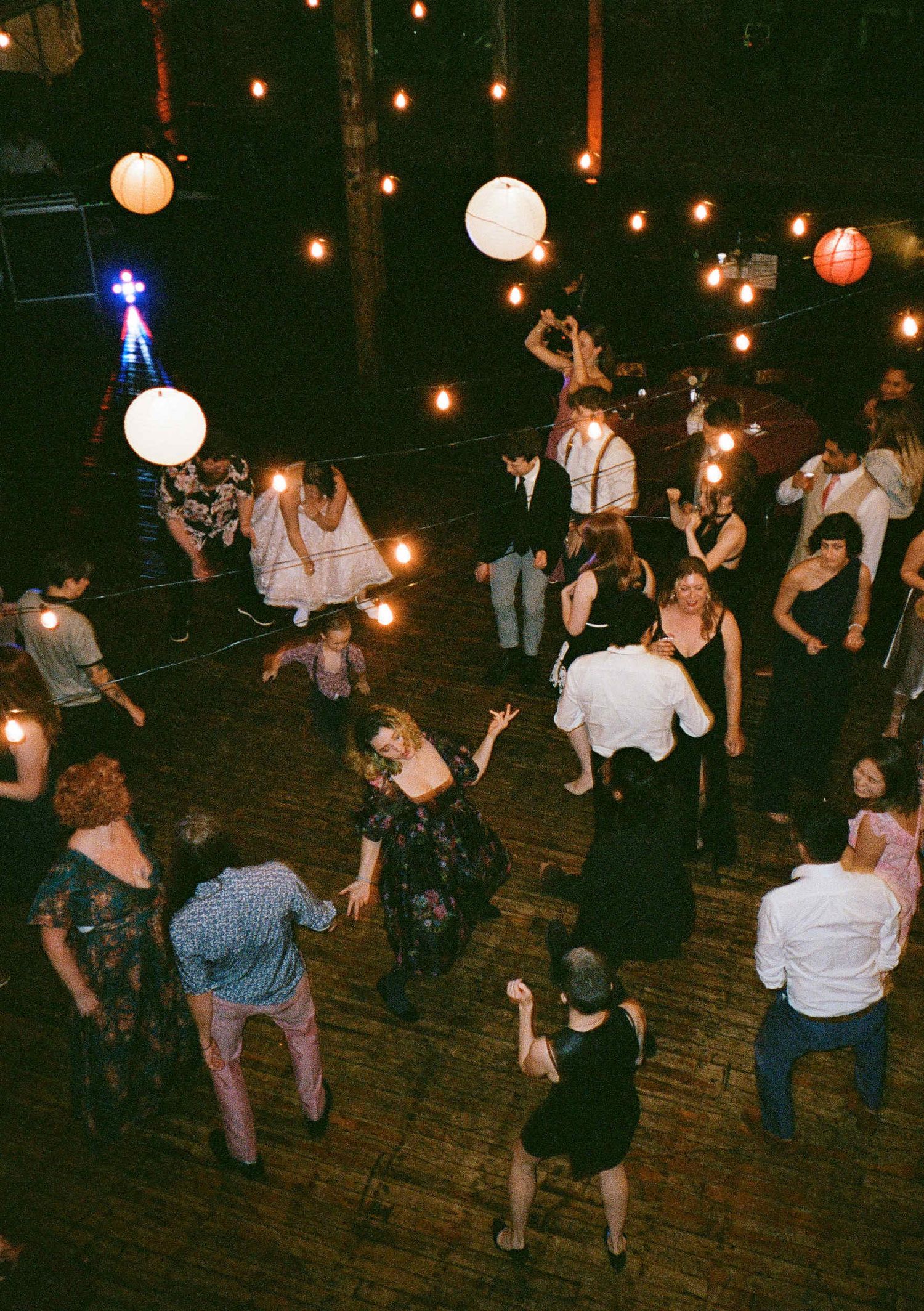 People dancing and celebrating at a dimly lit indoor venue with hanging paper lanterns and string lights on wooden floors.