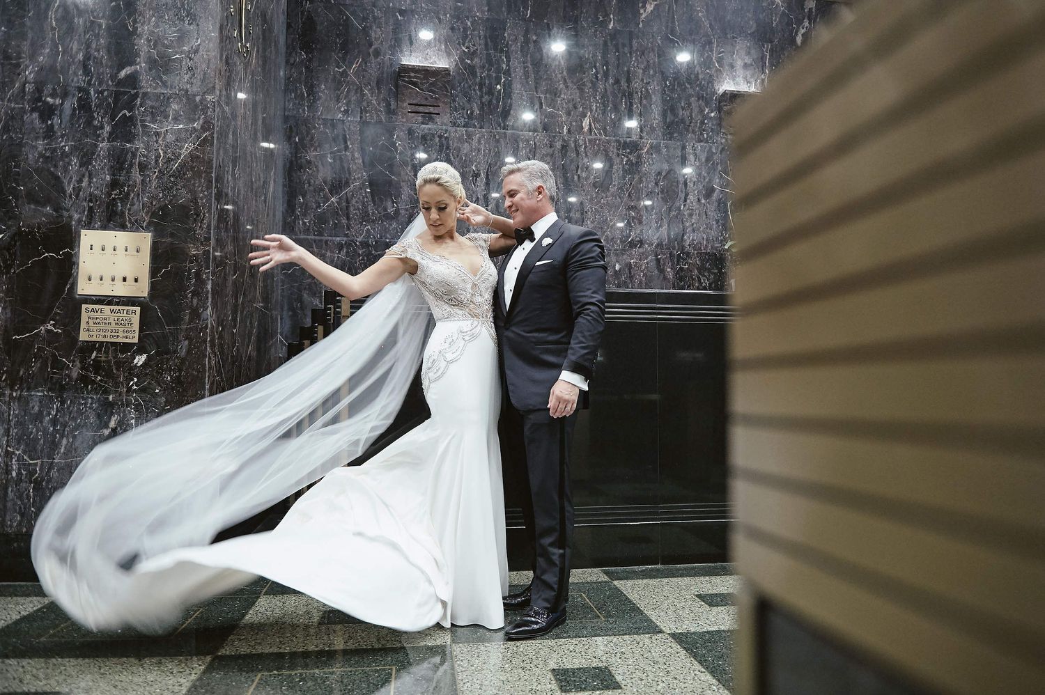 A newlywed couple in formal wedding attire dances together against a dark marble wall with a flowing white veil.